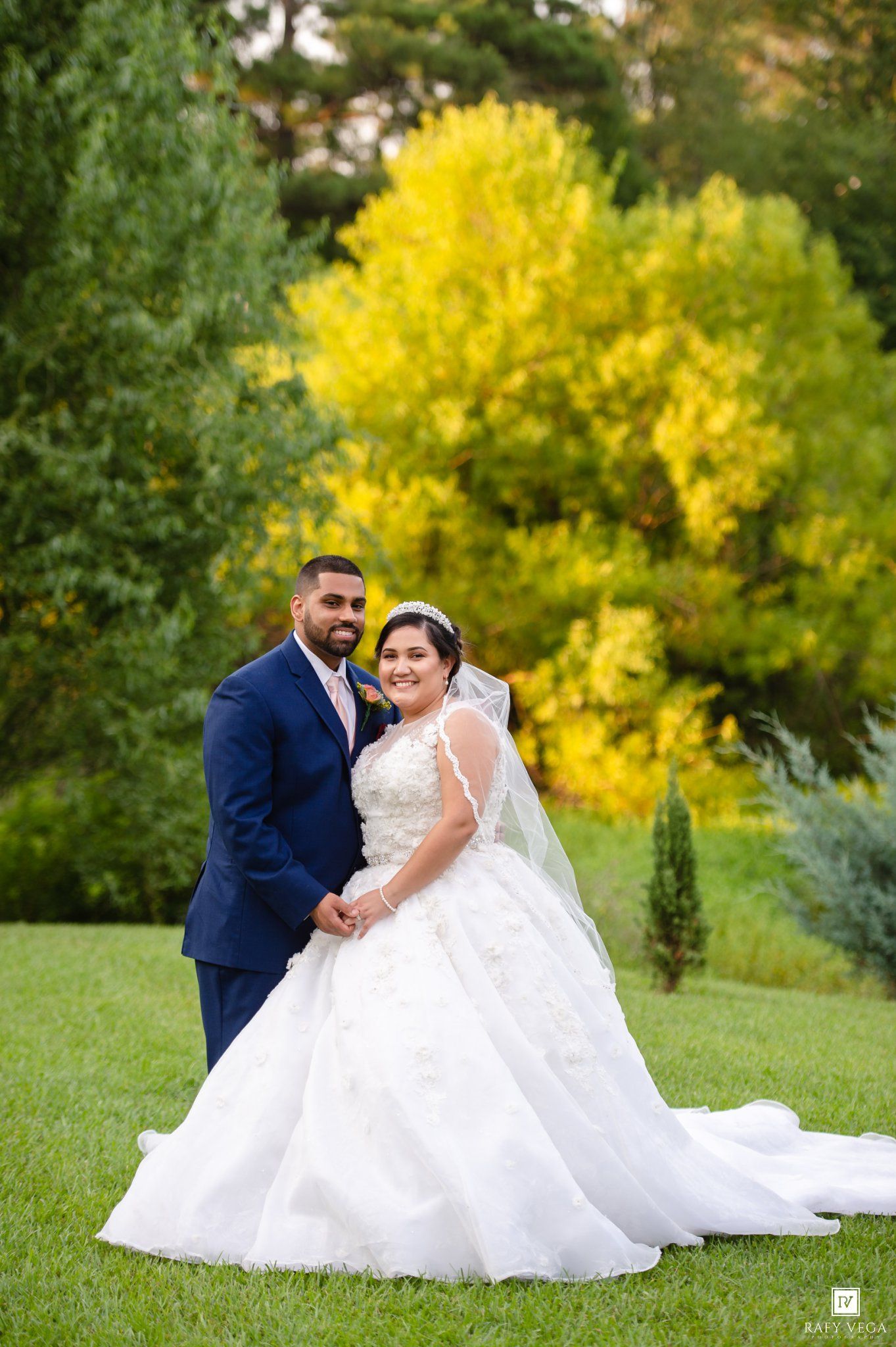 Wedding couple smiling outdoors among vibrant autumn trees, bride in white ball gown, groom in navy blue suit.