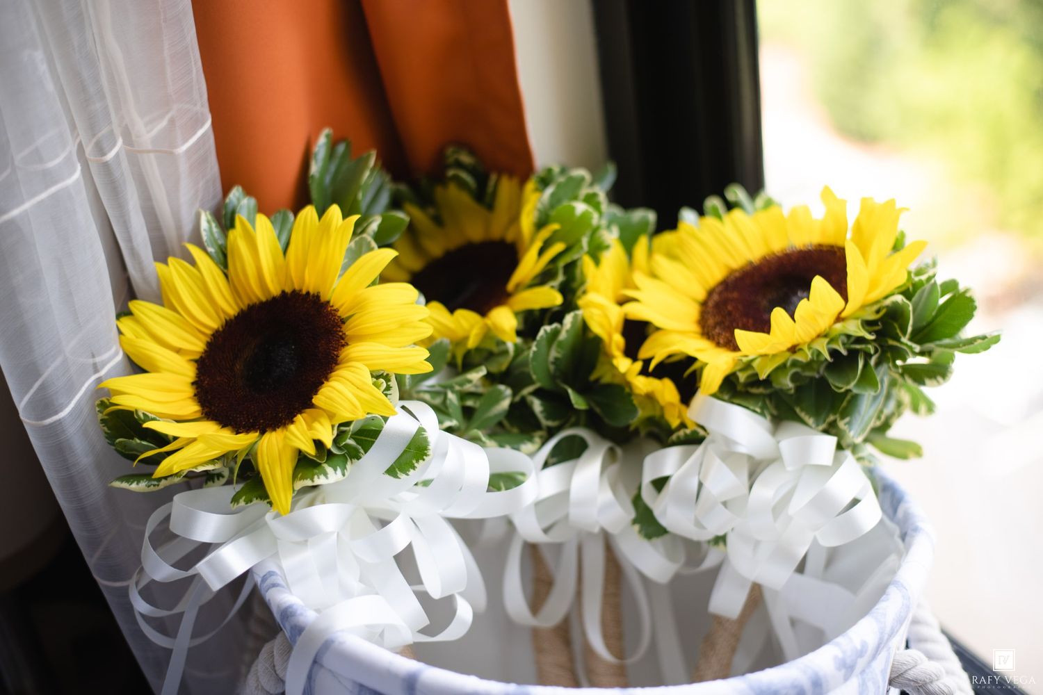 Two bright yellow sunflowers with white ribbon bows in a decorative basket, perfect for a wedding or special occasion.