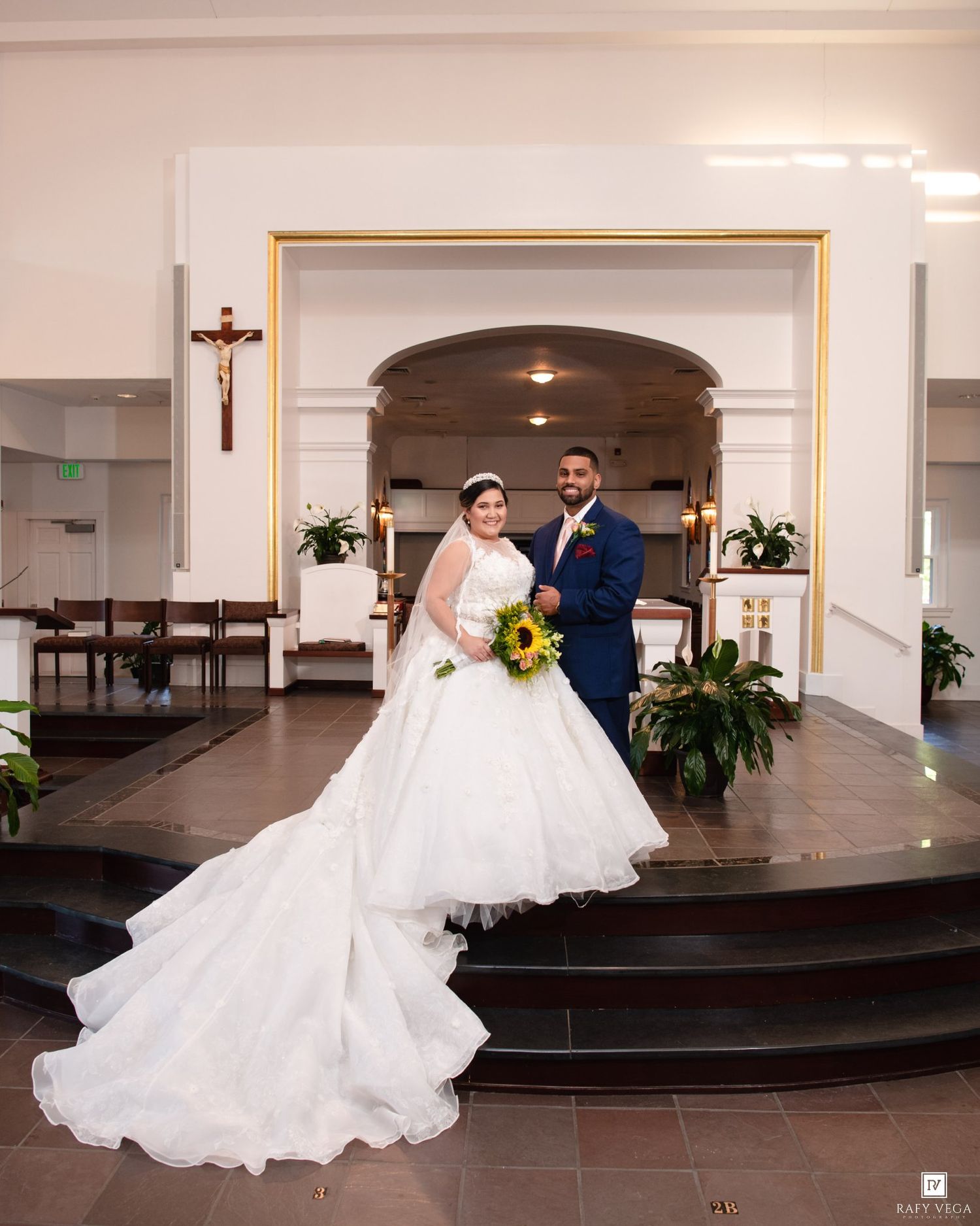 Bride in white ballgown and groom in navy suit pose with sunflower bouquet inside a Catholic church.