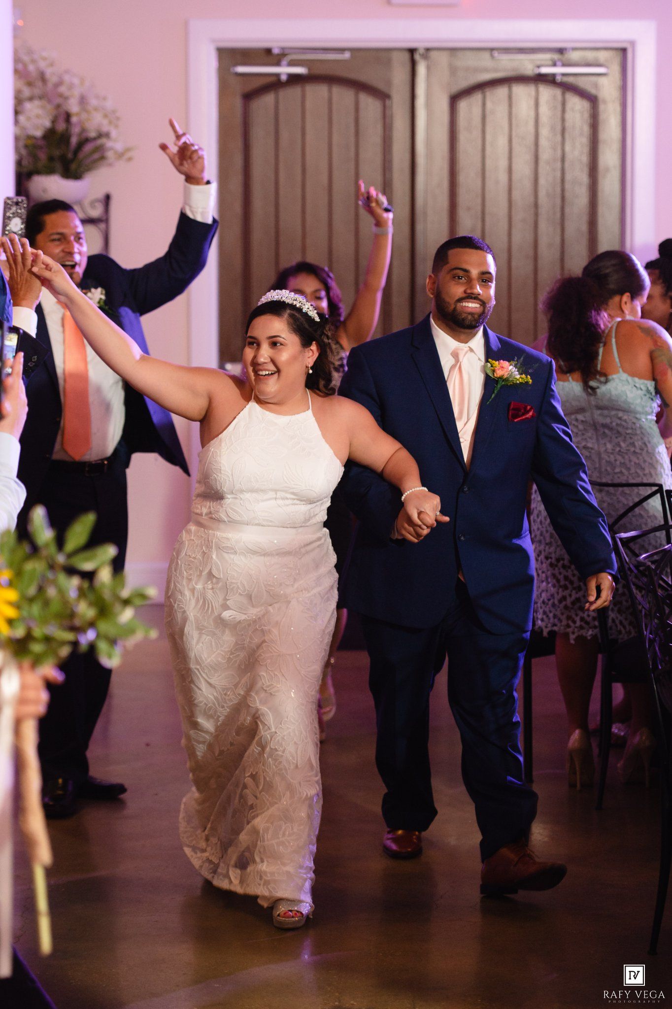 Bride and groom make joyful grand entrance at wedding reception, guests celebrating with raised arms in elegant venue.