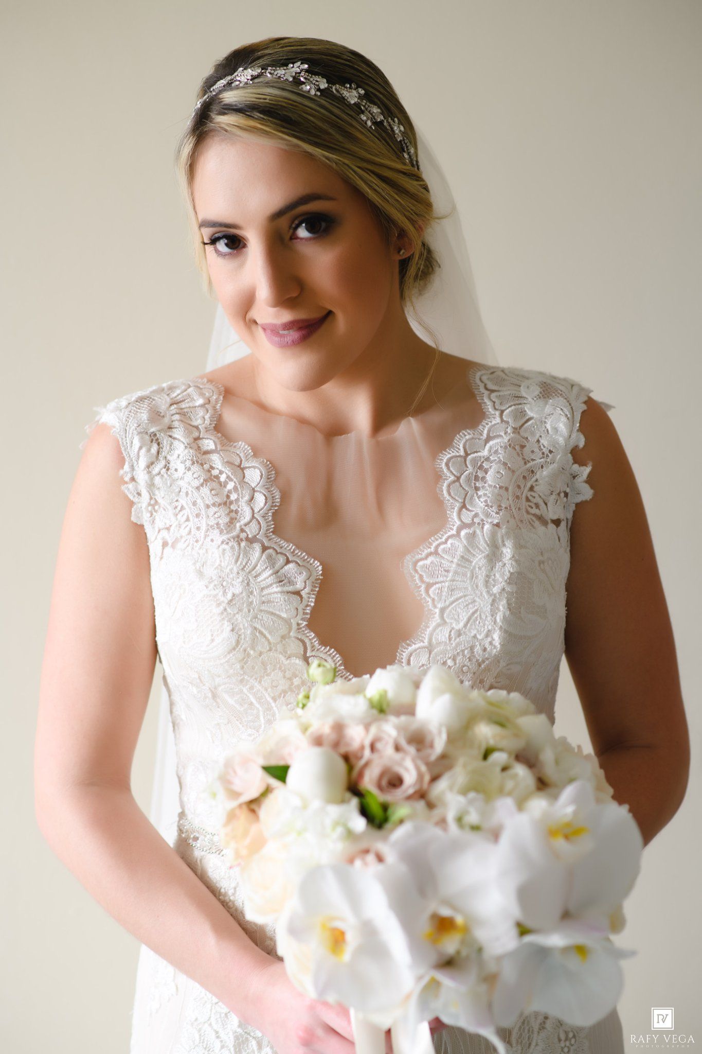 Bride in lace wedding dress holding white orchid bouquet with veil and floral headpiece, smiling softly.