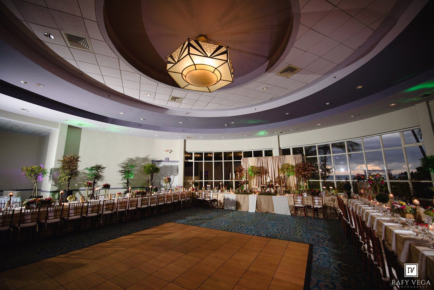 Elegant banquet hall with round ceiling, chandelier, long reception tables, floral centerpieces, and floor-to-ceiling windows at dusk.