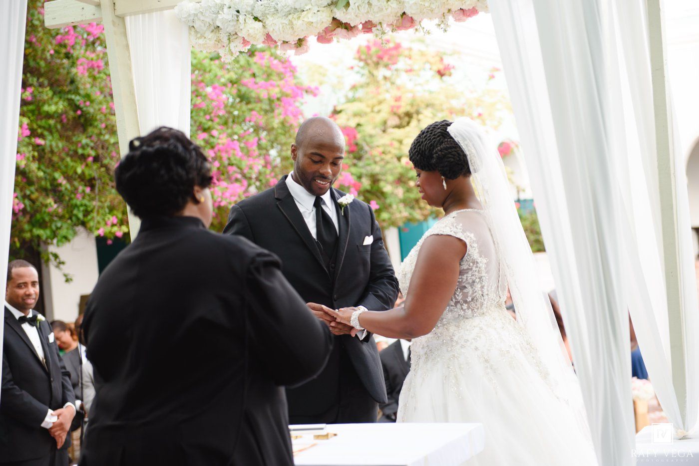 Bride and groom exchange vows under floral canopy with officiant at outdoor wedding ceremony.