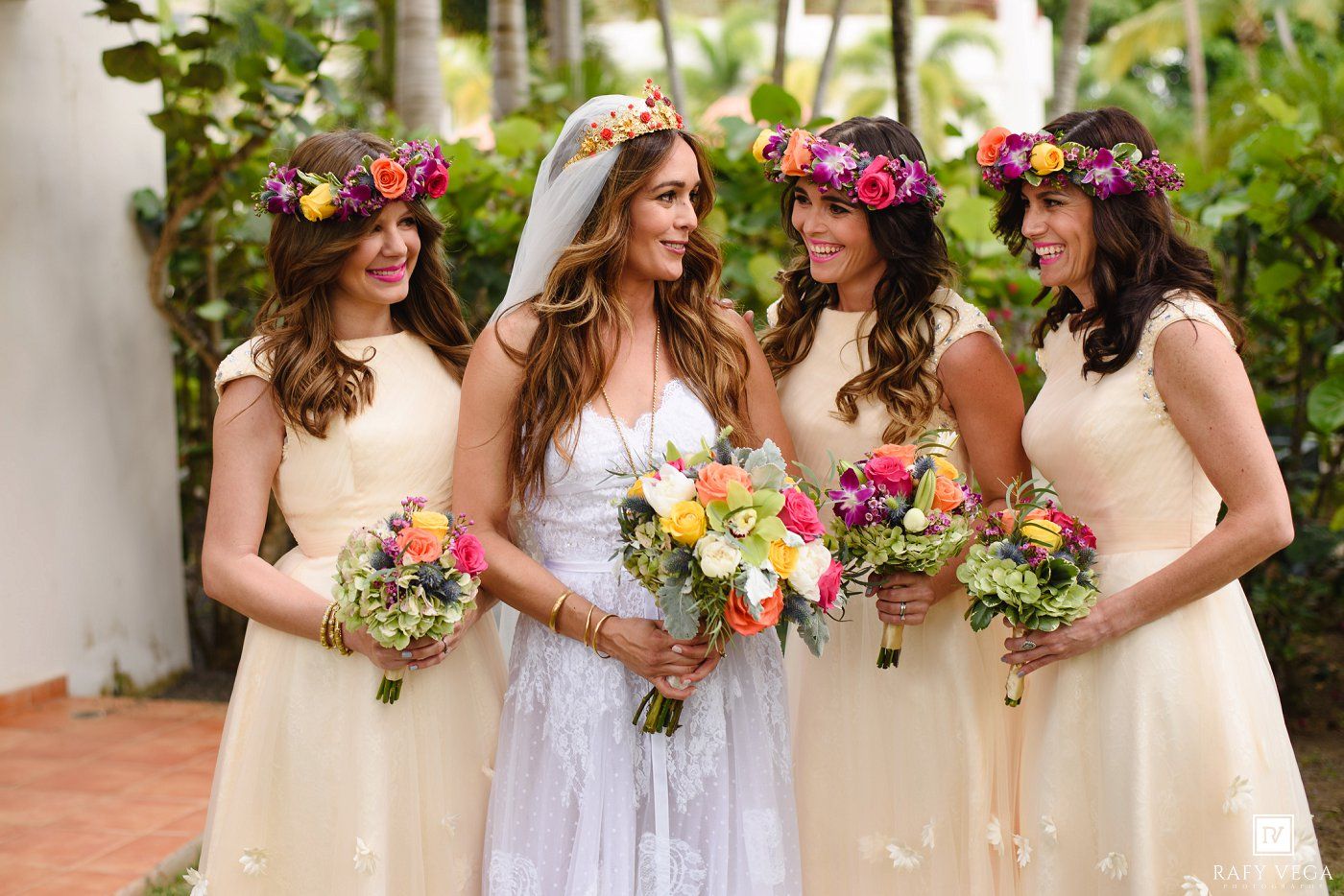 Bride in white gown with bridesmaids wearing cream dresses and colorful floral crowns holding vibrant bouquets outdoors.