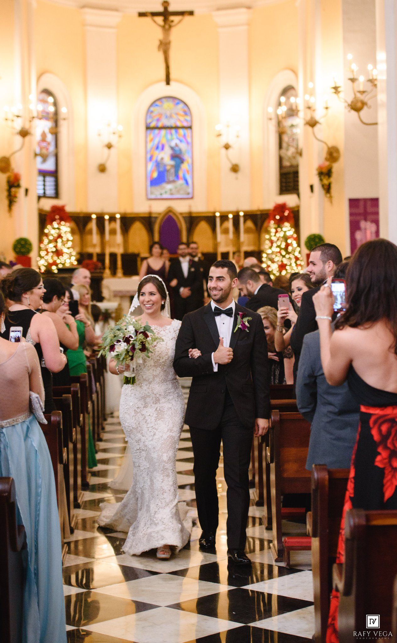 Bride and groom walk down church aisle after wedding ceremony, surrounded by guests, Christmas trees, and stained glass windows.