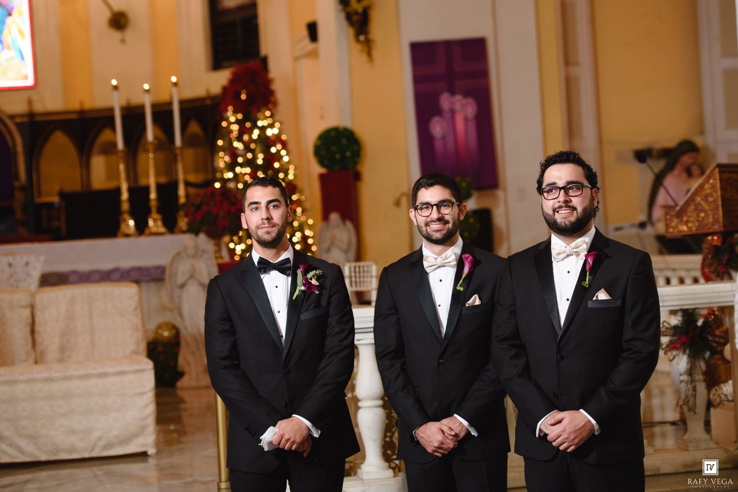 Three groomsmen in black tuxedos with floral boutonnieres standing at church altar during wedding ceremony.