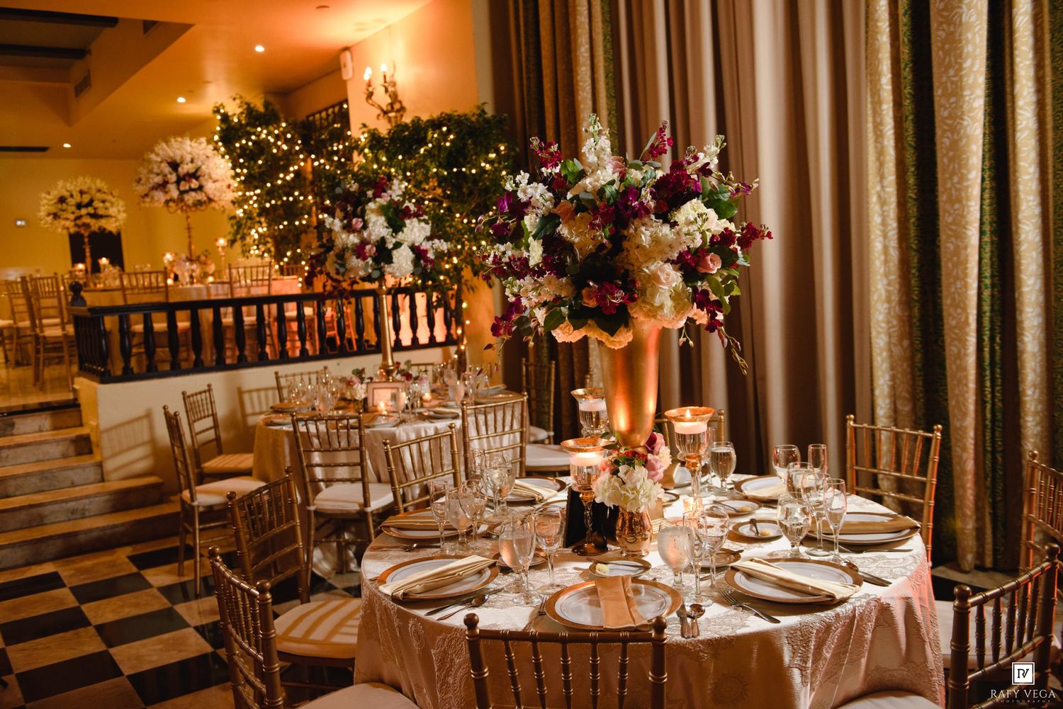 Elegant wedding reception with gold chiavari chairs, round tables with blush linens, and tall floral centerpieces in a warmly lit ballroom.