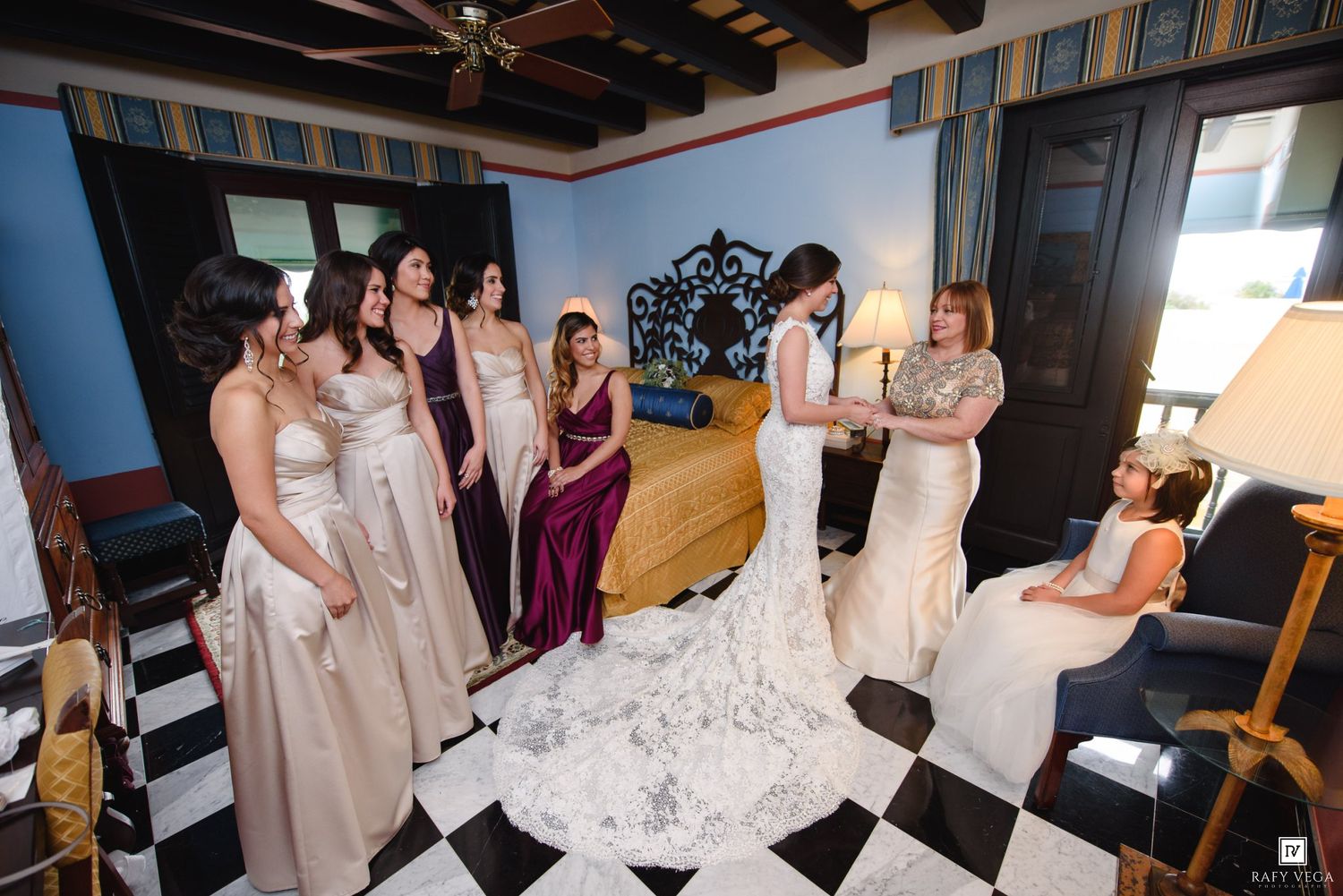 Bride in white lace gown surrounded by bridesmaids in elegant dresses in a decorative room with checkered floors.