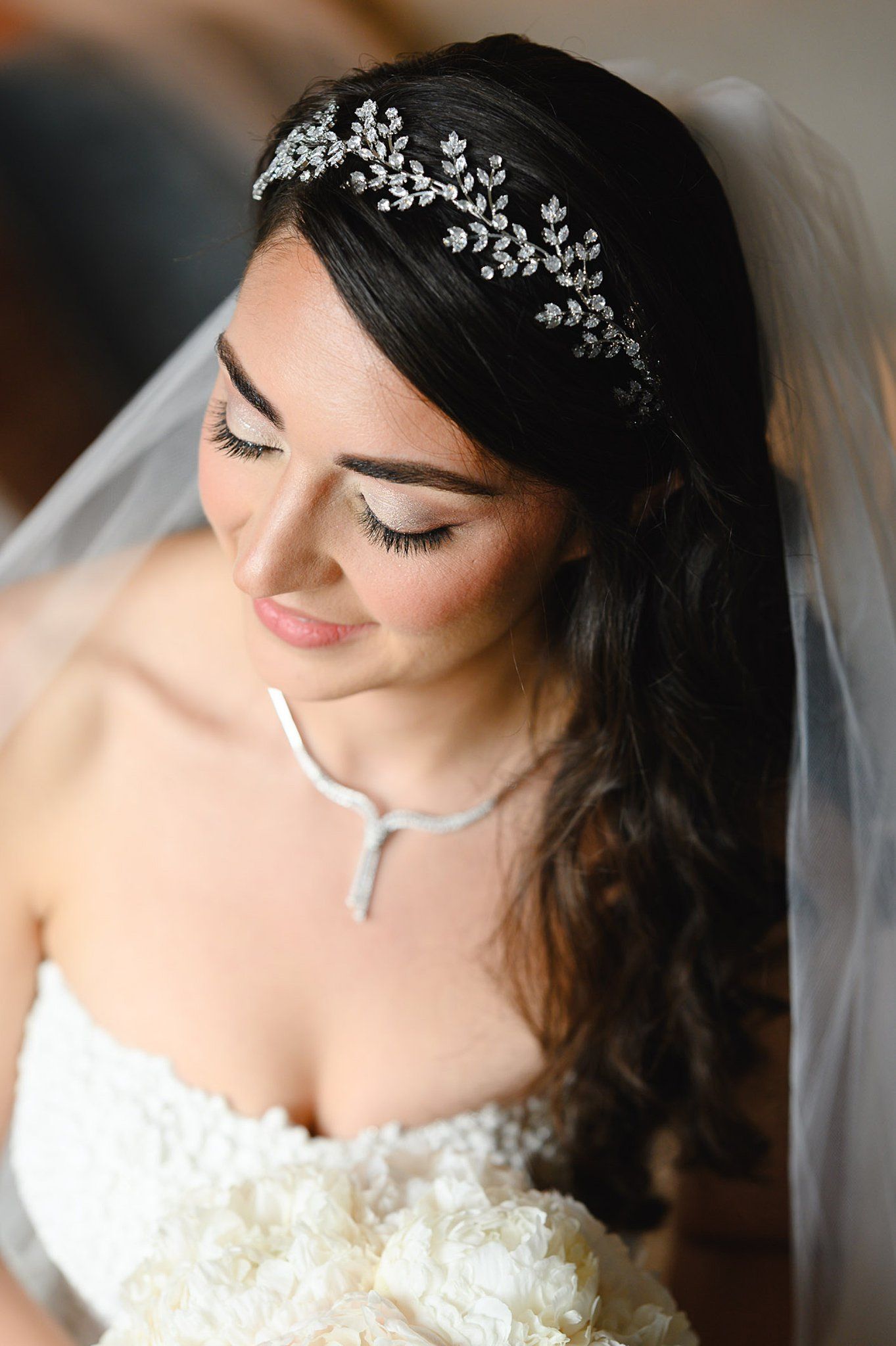 Bride wearing a crystal leaf headband with veil, natural makeup, and a white floral strapless gown, looking down gracefully.