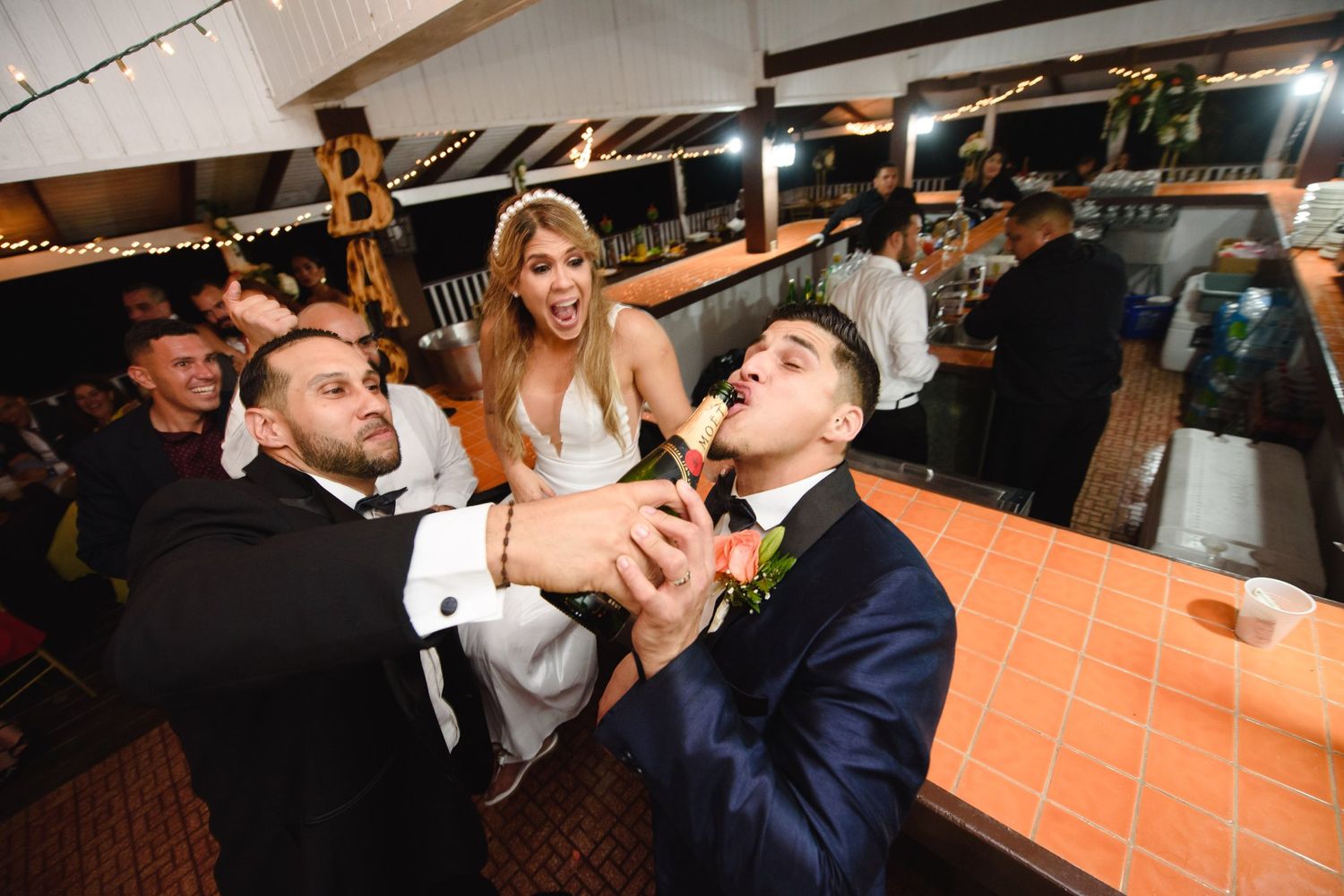 Bride and groom celebrate at wedding reception as guest pours drink into groom's mouth at indoor venue.
