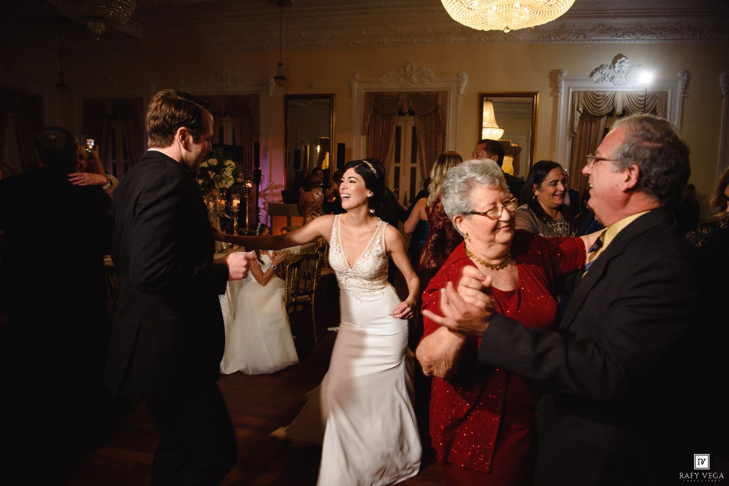 Bride in white gown dances joyfully with guests including elderly woman in red dress at elegant wedding reception.