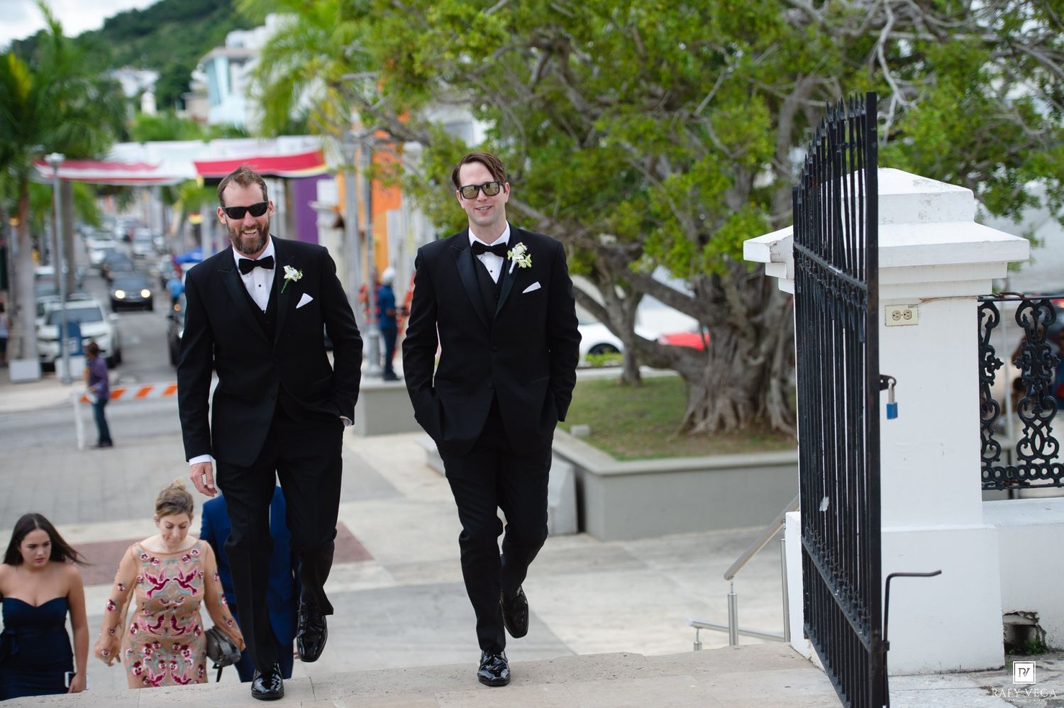 Two groomsmen in black tuxedos walking down a sunny sidewalk toward a wedding venue.