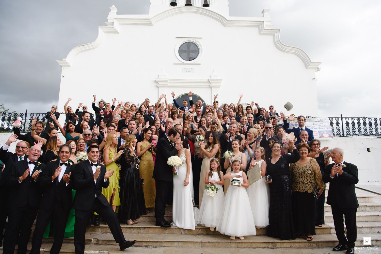 Large wedding party celebrating on white church steps with bride, groom, guests cheering outdoors.