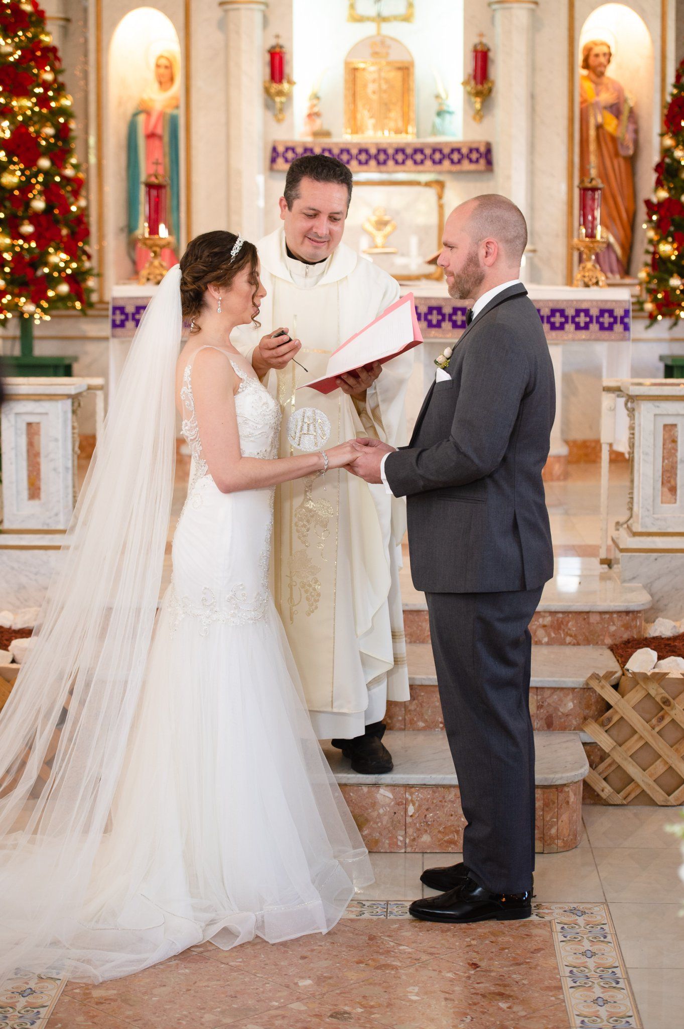 Bride and groom exchange vows before a priest at a decorated Catholic church altar during a Christmas wedding ceremony.