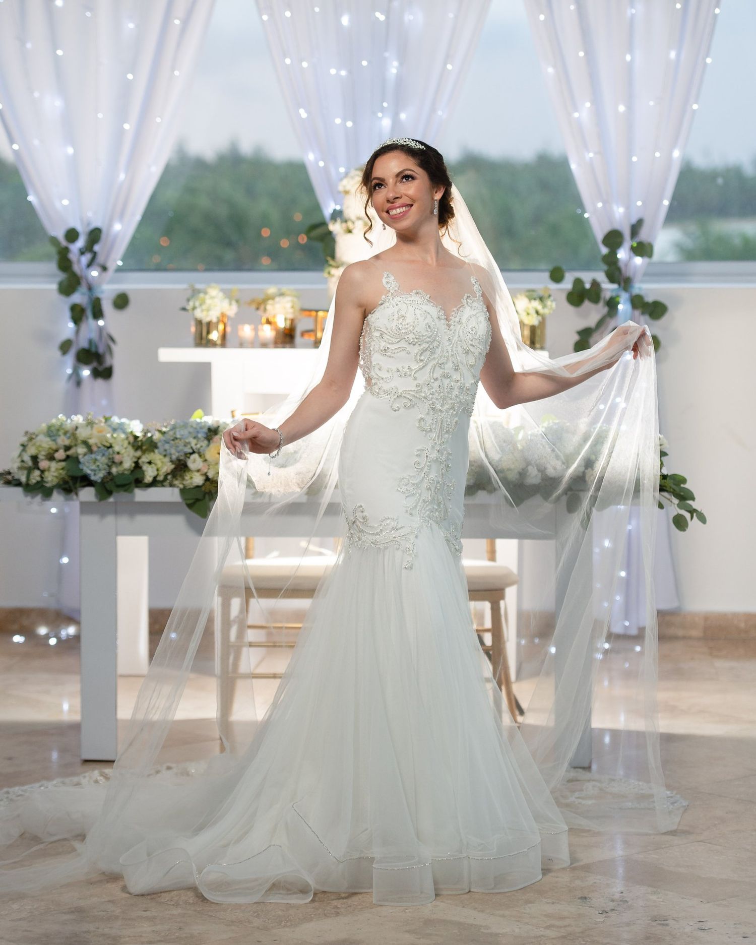 Bride in fitted lace wedding gown spreading her veil at decorated ceremony venue with twinkling lights and greenery.