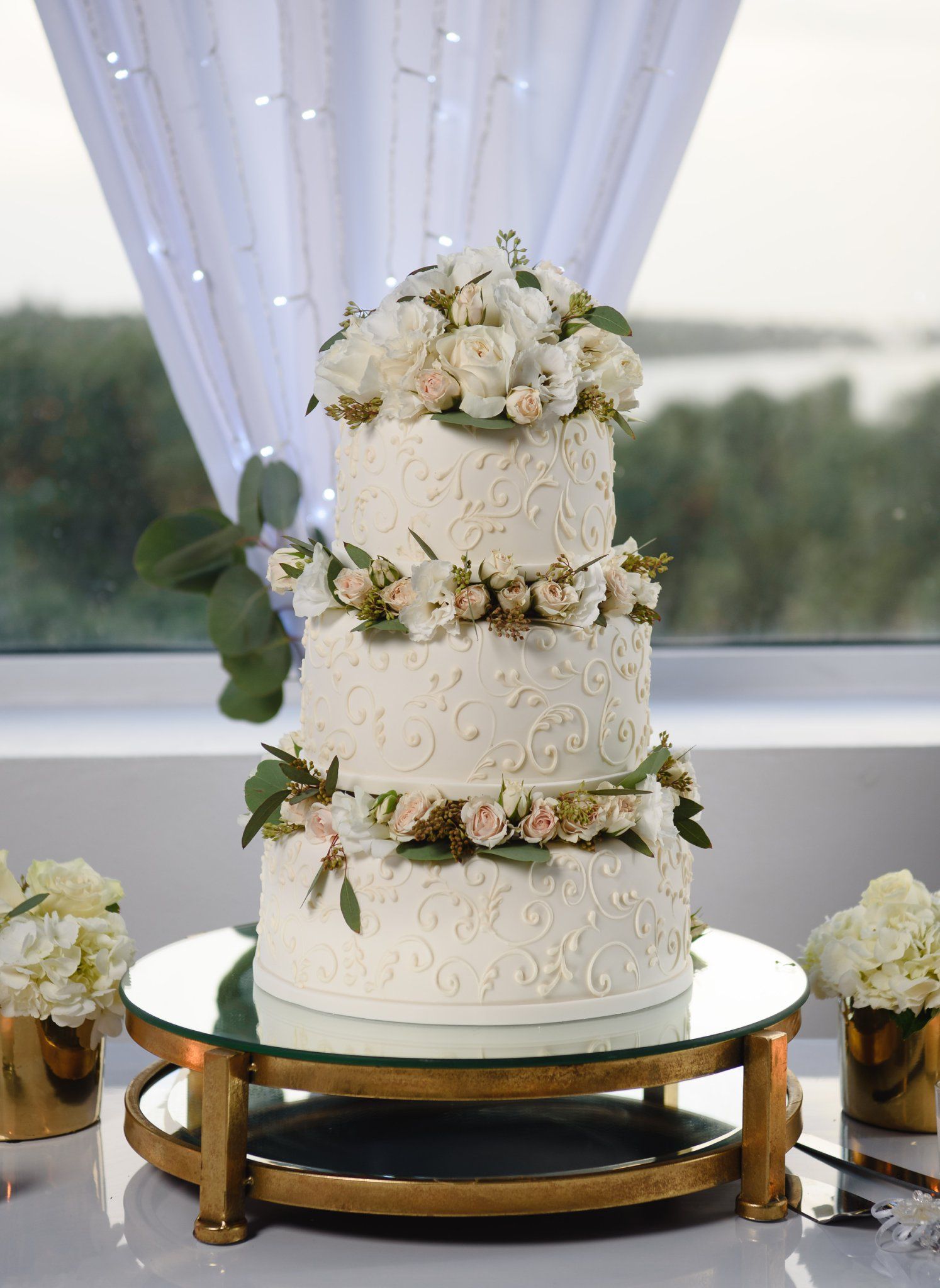 Elegant three-tier white wedding cake with floral decorations and scroll details on a gold mirrored stand.