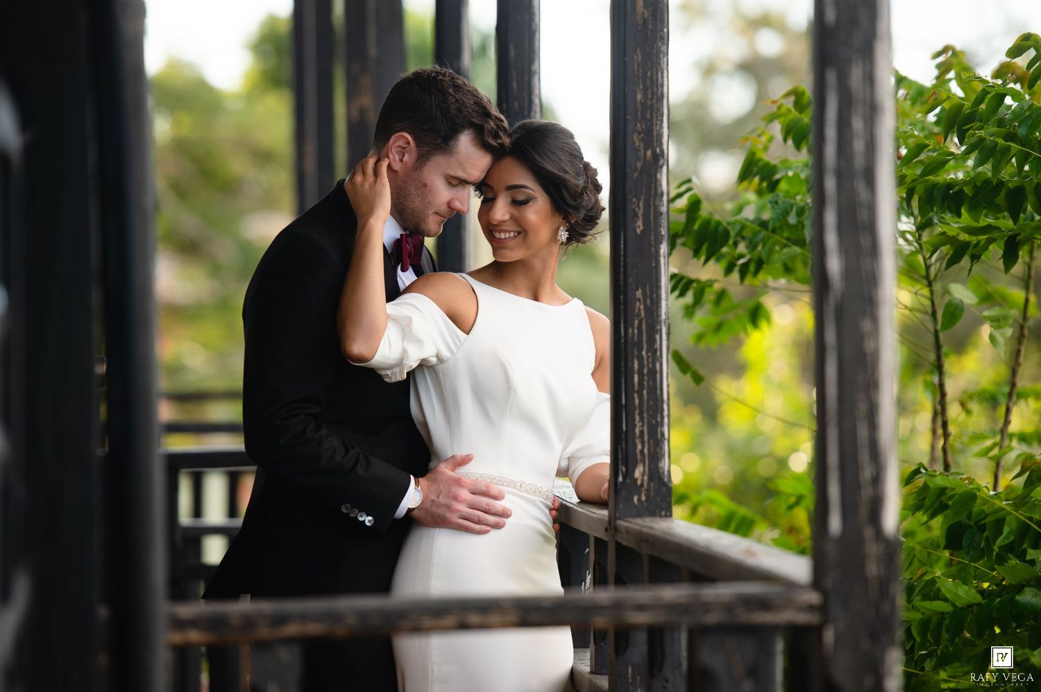 Bride and groom share a romantic embrace on a wooden balcony surrounded by lush greenery.