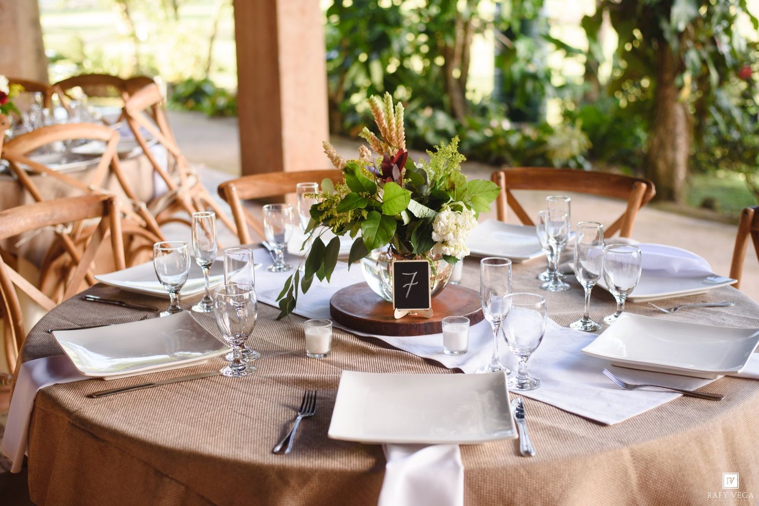 Elegant rustic wedding reception table set with burlap linens, white plates, crystal glasses, and floral centerpiece.