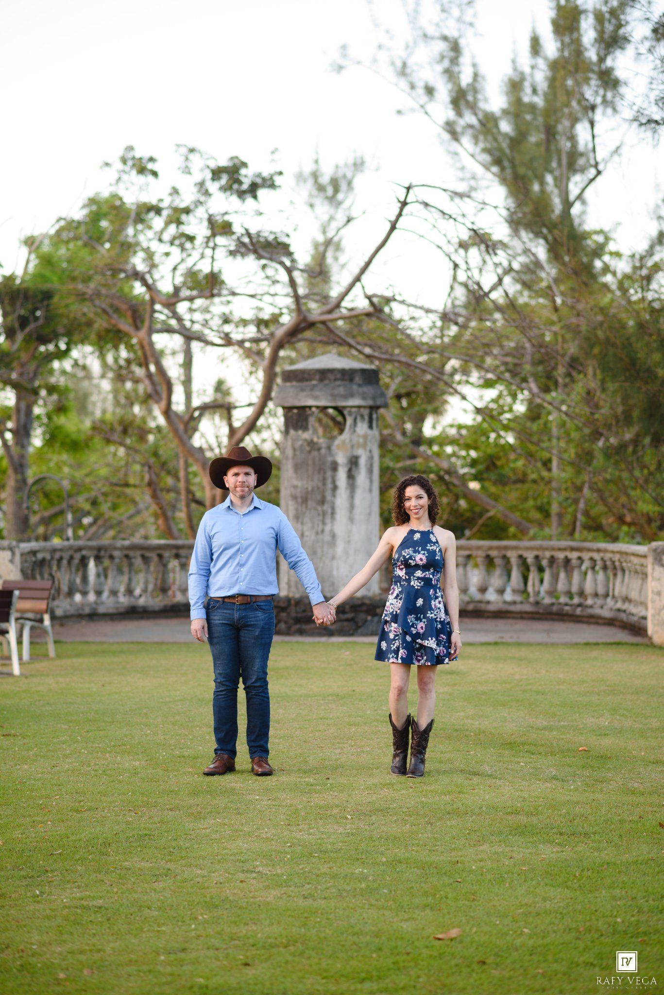 Couple holding hands on green lawn, man in cowboy hat and blue shirt, woman in floral dress and boots.