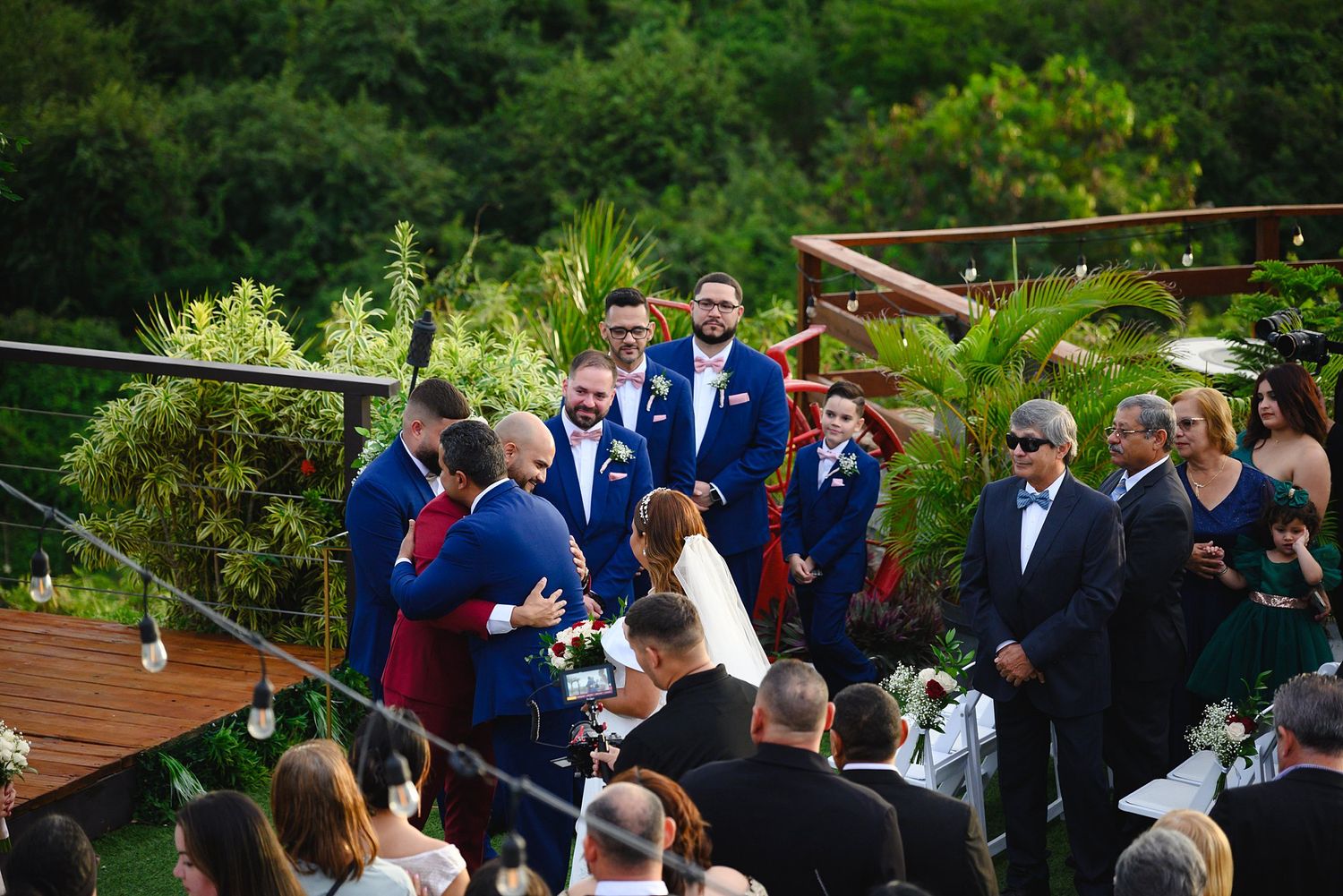 Outdoor wedding ceremony with groom and groomsmen in blue suits on a wooden deck surrounded by lush greenery.