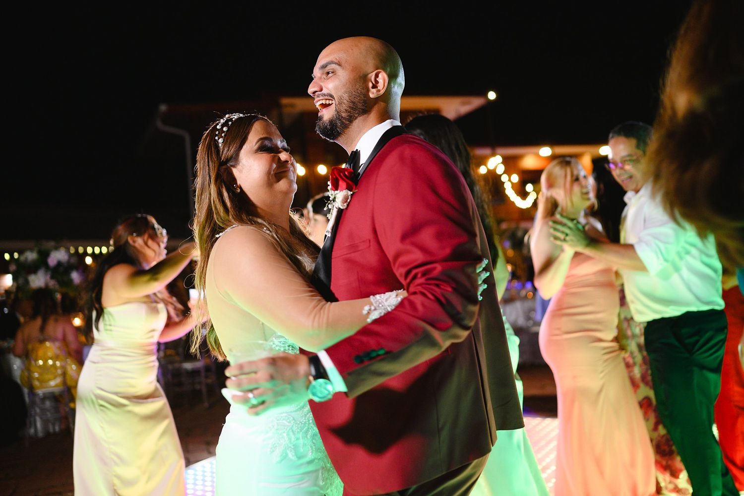 Bride and groom share first dance at outdoor wedding reception with guests dancing in background at night.