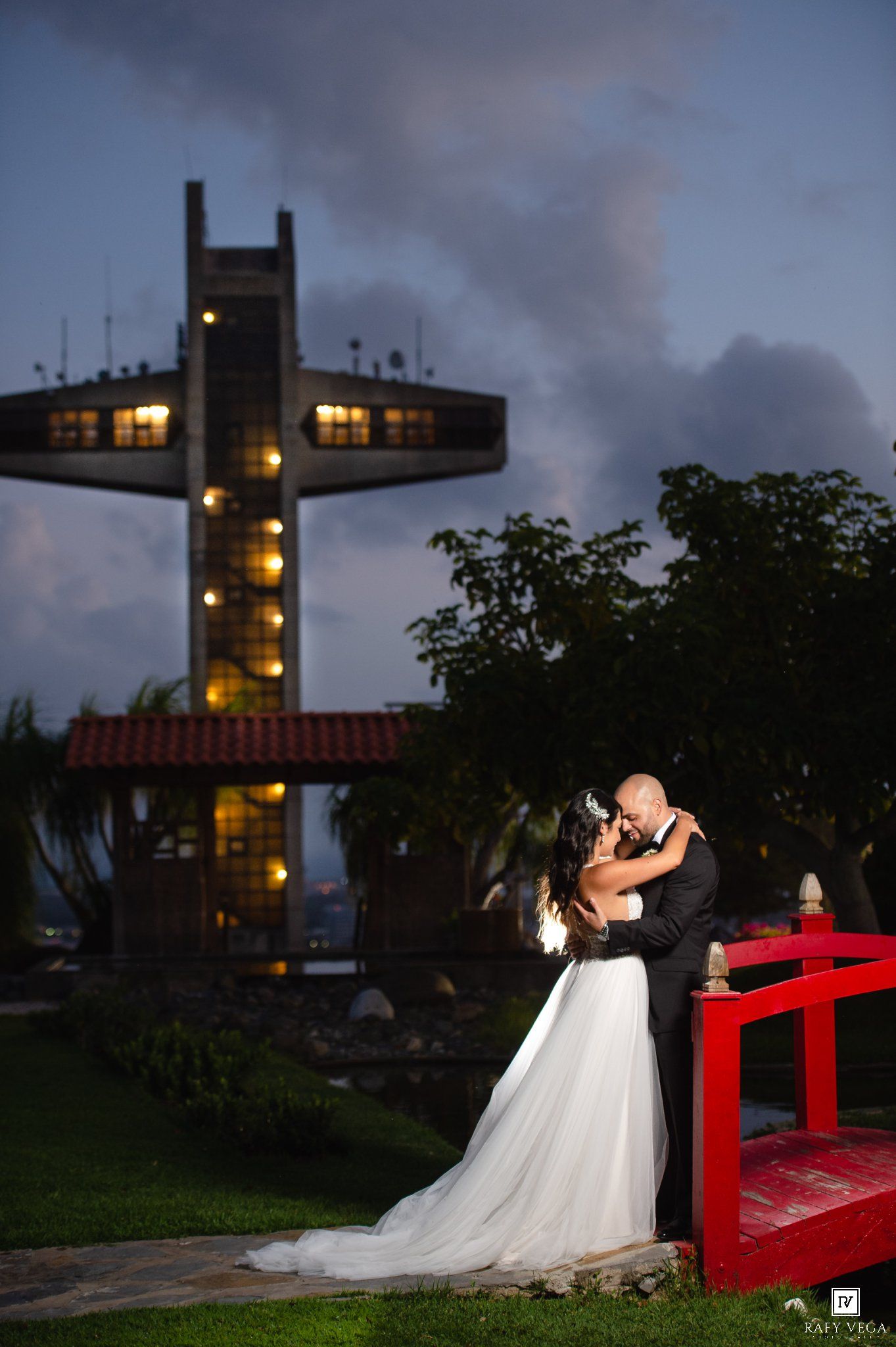Bride and groom share romantic kiss at dusk near illuminated cross tower and red bridge at outdoor wedding venue.