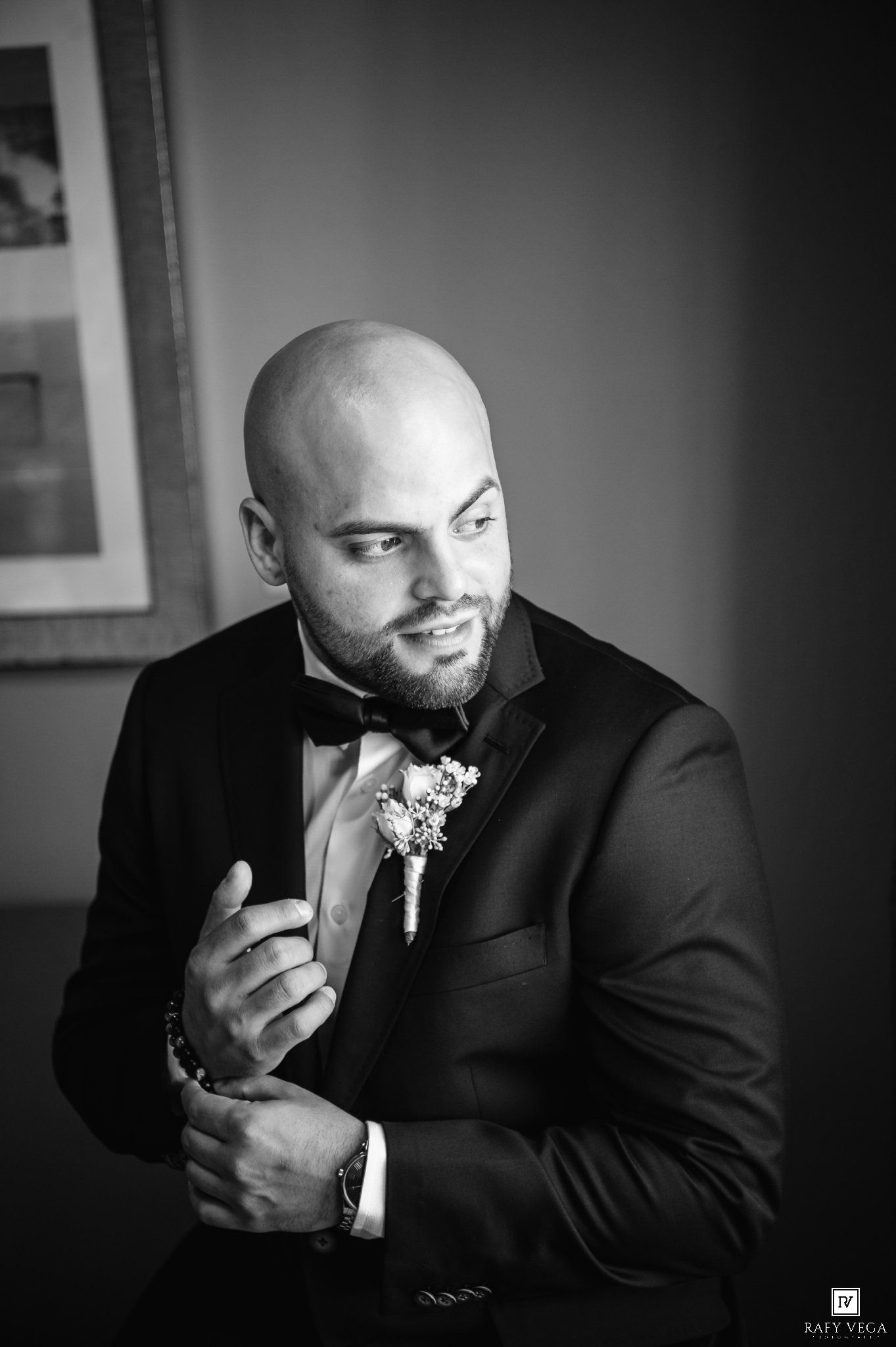 Black and white portrait of a bald groom in a tuxedo with bow tie and boutonniere, adjusting his cufflinks.
