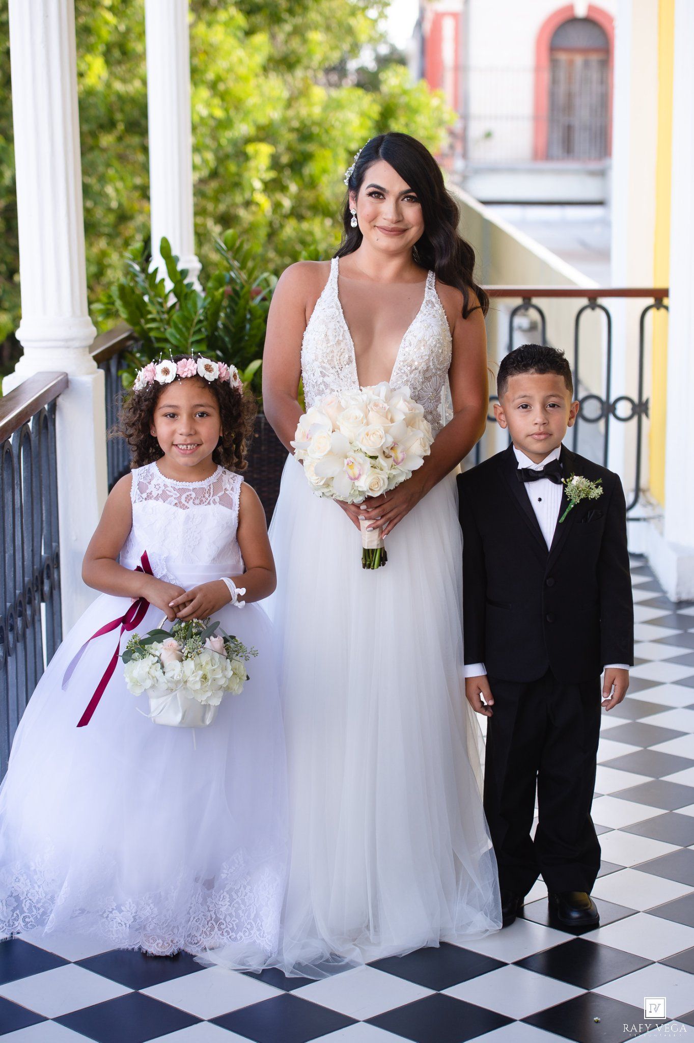 Bride in white tulle gown poses with flower girl and ring bearer on checkered balcony floor.