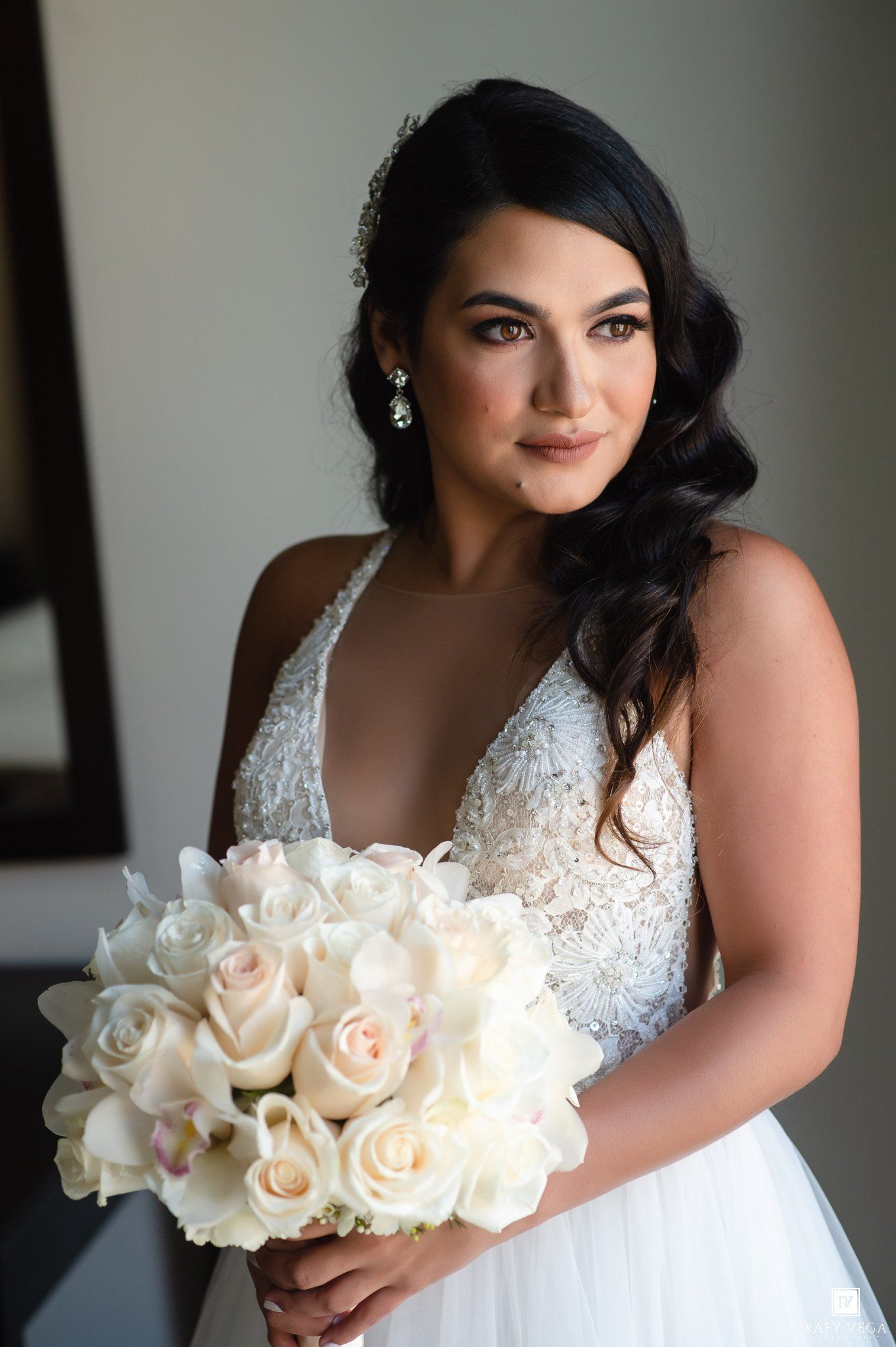 Bride in lace wedding gown holds white rose bouquet, wearing pearl earrings and elegant hair accessories.