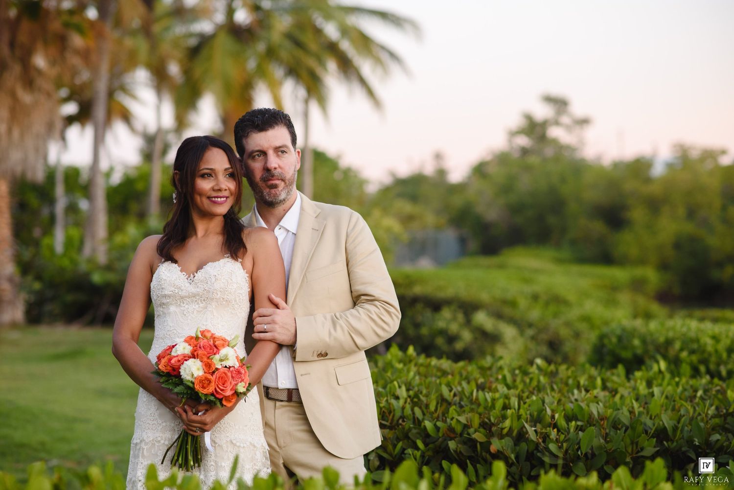 Smiling bride and groom pose outdoors at tropical wedding venue with lush greenery and palm trees.