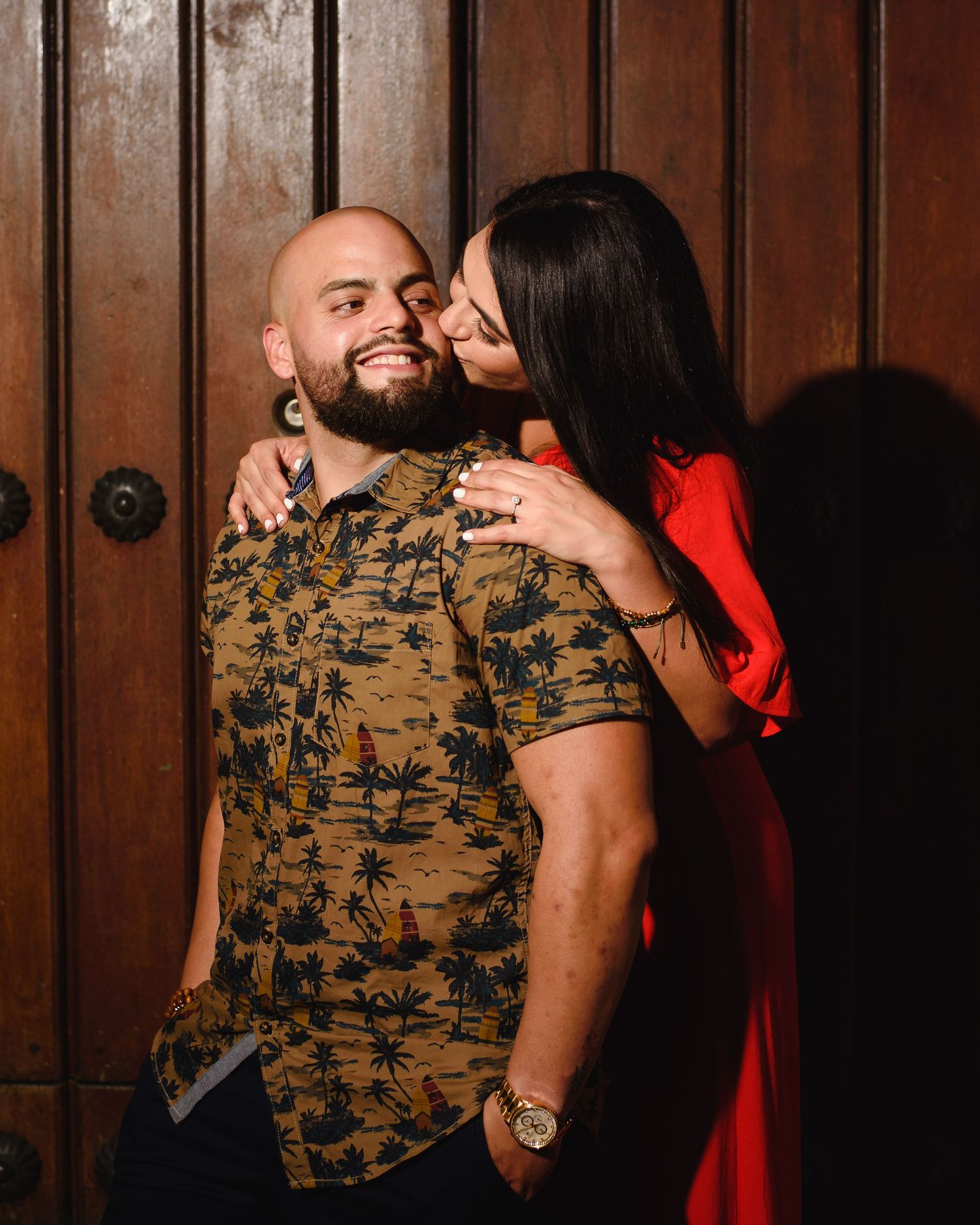 A smiling bald man with a beard in a tropical shirt receives a kiss on the cheek from a woman in a red dress.
