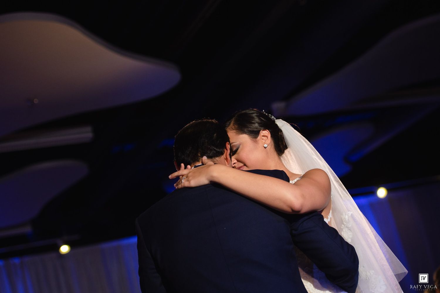 Bride and groom share an emotional first dance embrace at their wedding reception under blue ambient lighting.