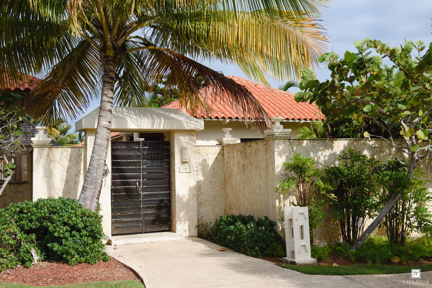 Tropical villa entrance with red-tiled roof, ornate wooden gate, stucco walls, and lush palm trees in a sunny Caribbean setting.