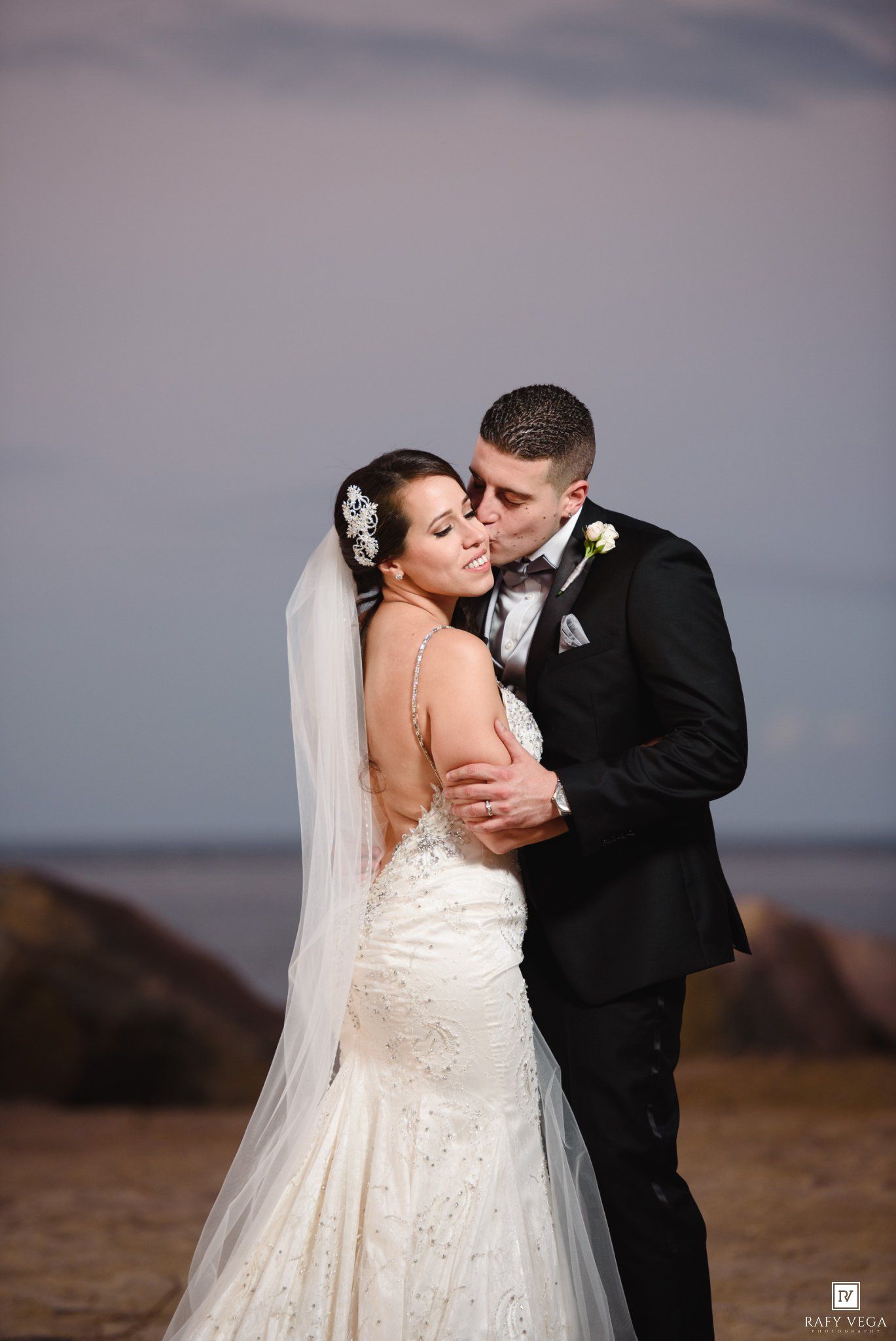 A bride and groom share a romantic embrace on a beach at sunset, she in a beaded mermaid gown and he in a black tuxedo.
