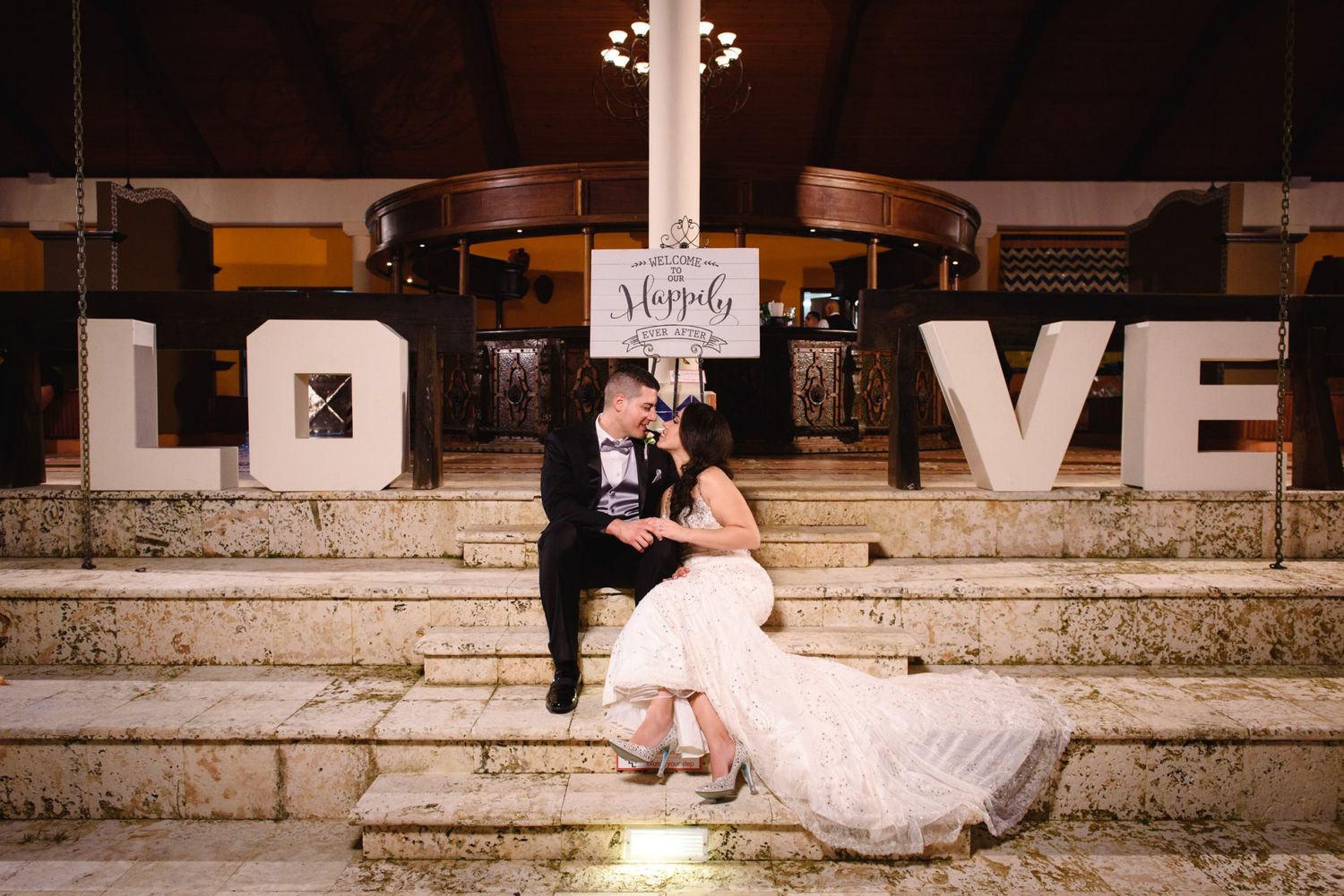 Bride and groom sit romantically on stone steps between large LOVE letters at an elegant evening wedding reception.