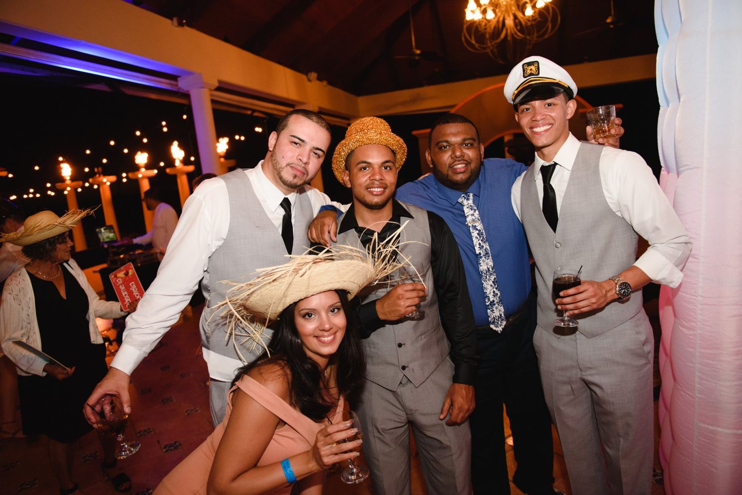 Group of five friends posing together at a festive indoor party, wearing hats and formal attire.