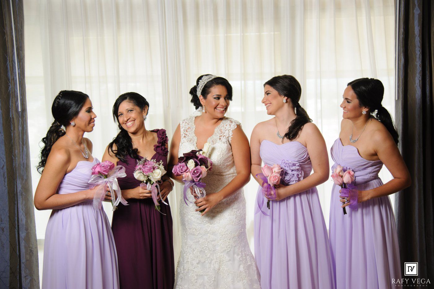 Bride in lace gown stands with four bridesmaids in lavender dresses holding pink and purple bouquets.