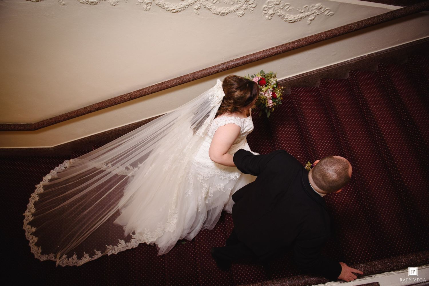 Bride in white gown and veil descending elegant carpeted staircase with groom, aerial view at wedding venue.