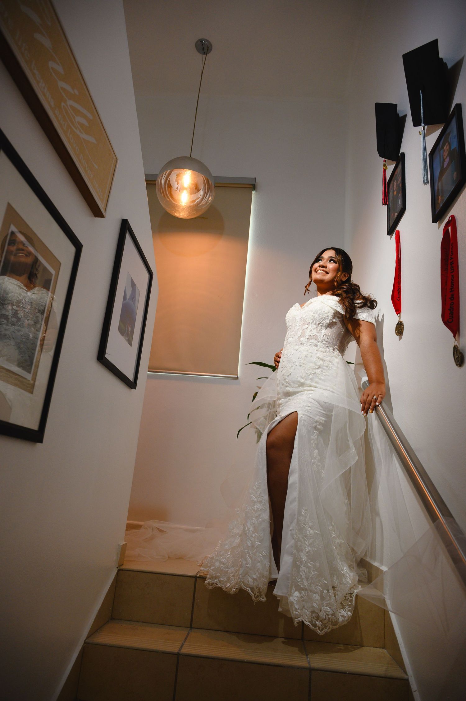 Bride in lace wedding gown with slit descends staircase, smiling joyfully under warm pendant light.