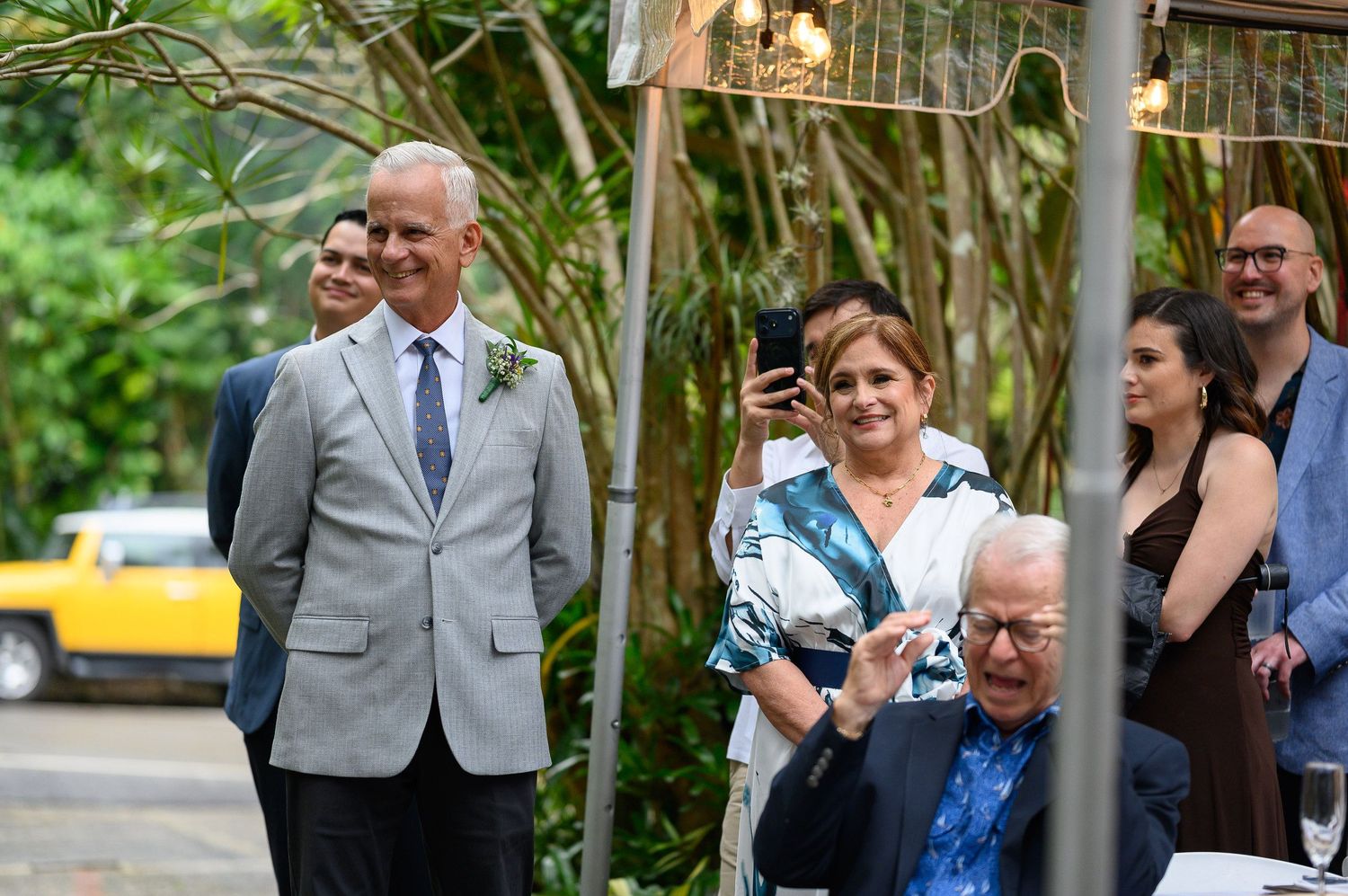 Elegant outdoor gathering with guests in formal attire, including a man in a gray suit and tie, smiling at a garden event.