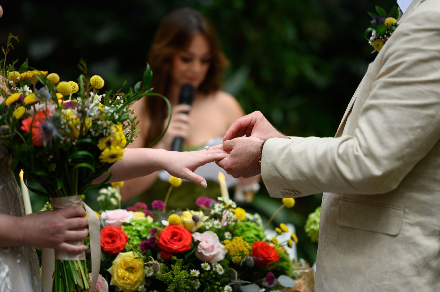 Couple exchanging rings during outdoor wedding ceremony with colorful floral bouquets and officiant in background.