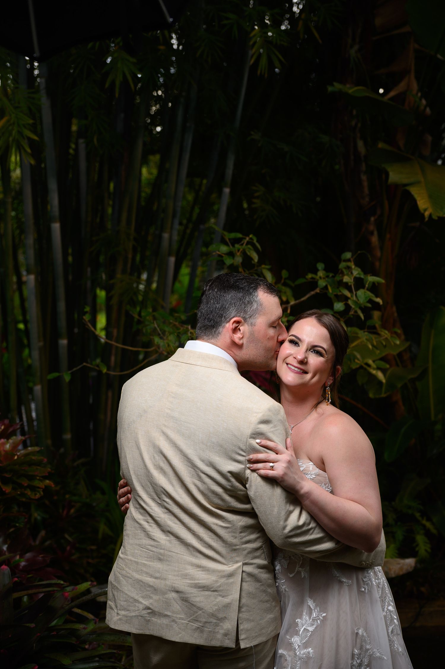 A bride and groom share a romantic embrace in a lush tropical garden, the groom kissing her cheek as she smiles.