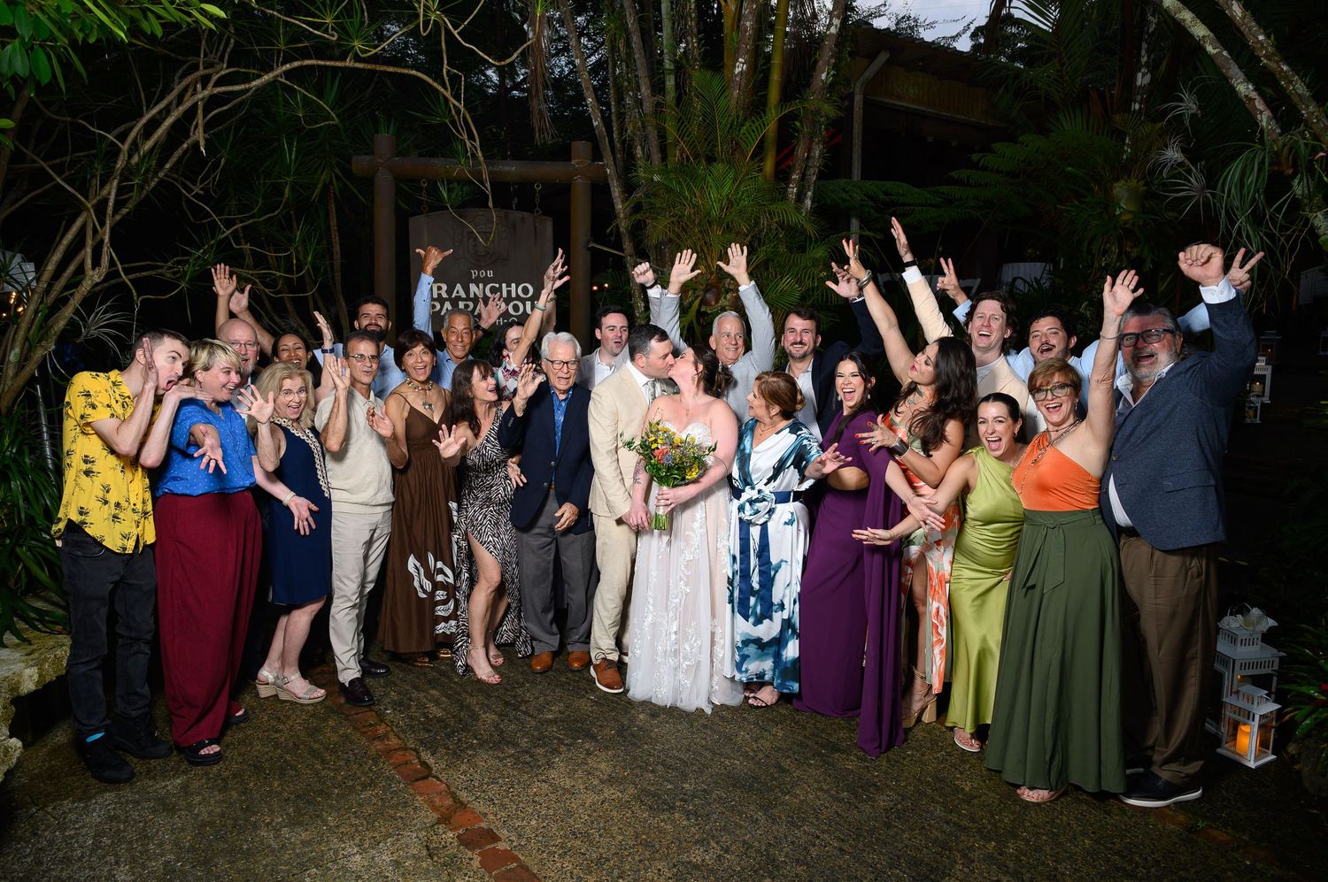 A joyful wedding group photo with guests cheering outdoors at a tropical garden venue at night.