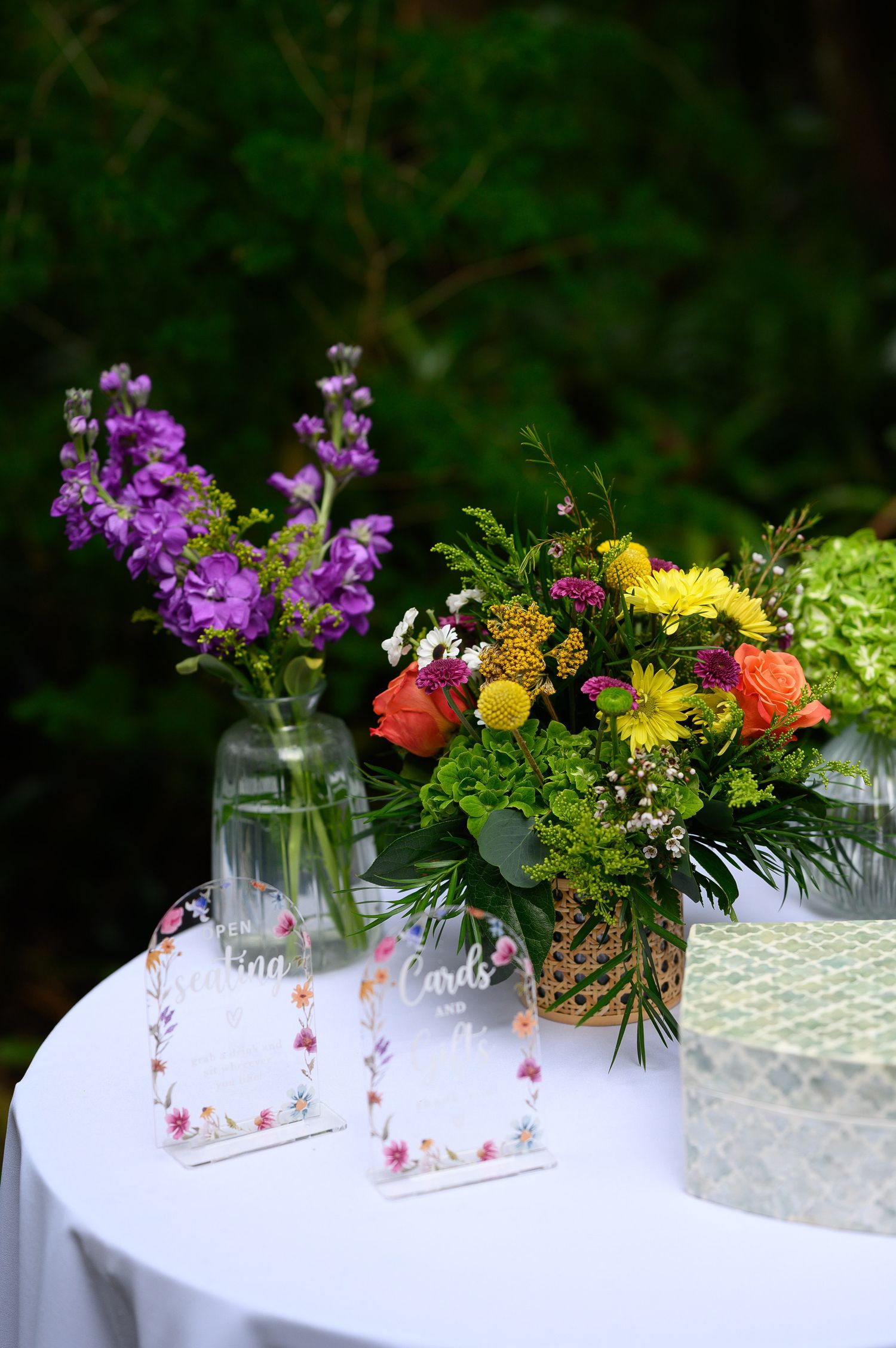 Colorful floral arrangements on a white table with wedding cards and gift box at an outdoor garden event.