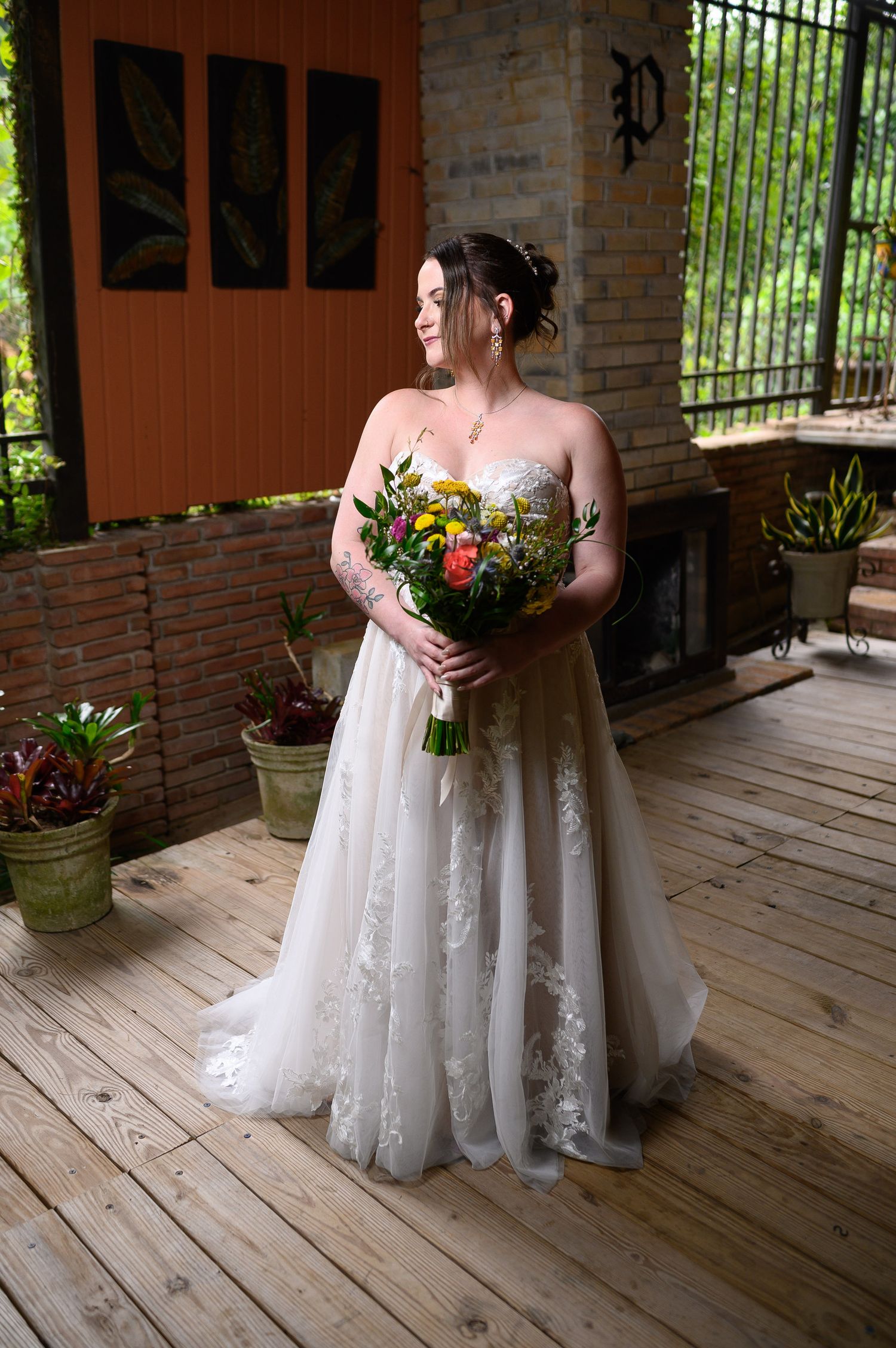 Bride in strapless lace wedding gown holding colorful bouquet on wooden deck with brick backdrop.