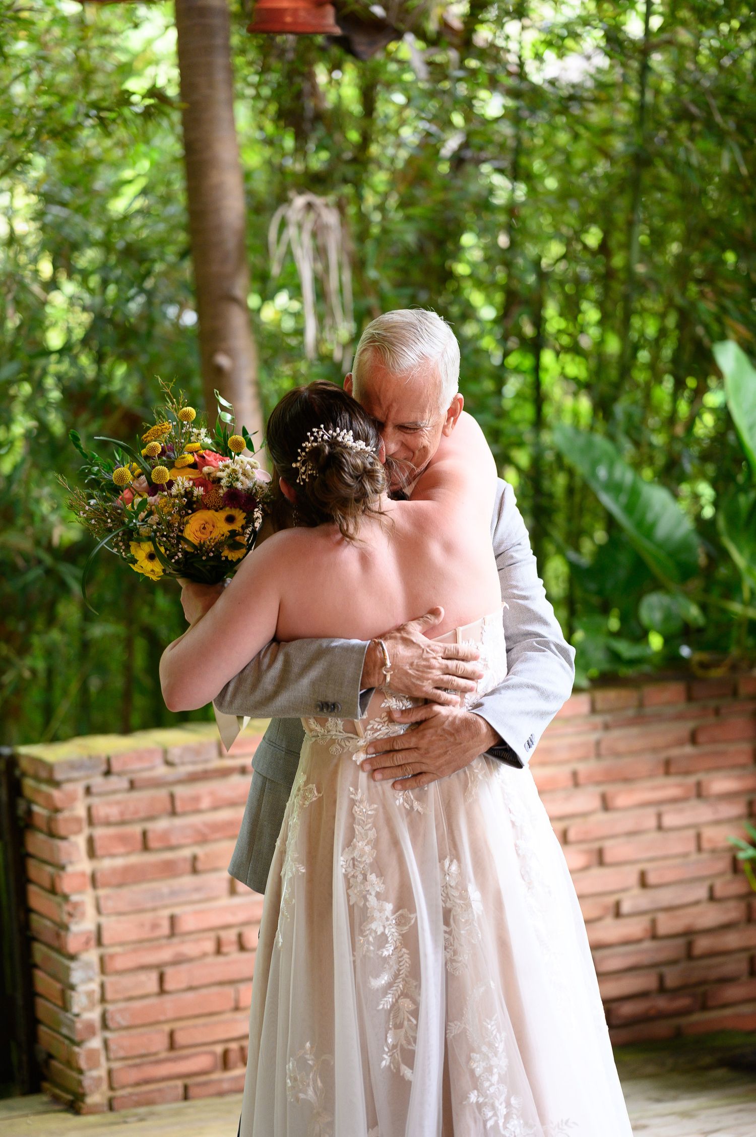 Bride in lace gown hugging older man outdoors, holding yellow floral bouquet at a garden wedding ceremony.