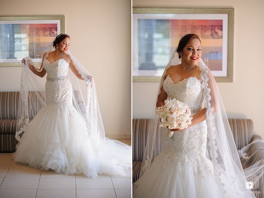 Bride in a strapless mermaid wedding gown with lace details holds her veil and bouquet in a bright room.