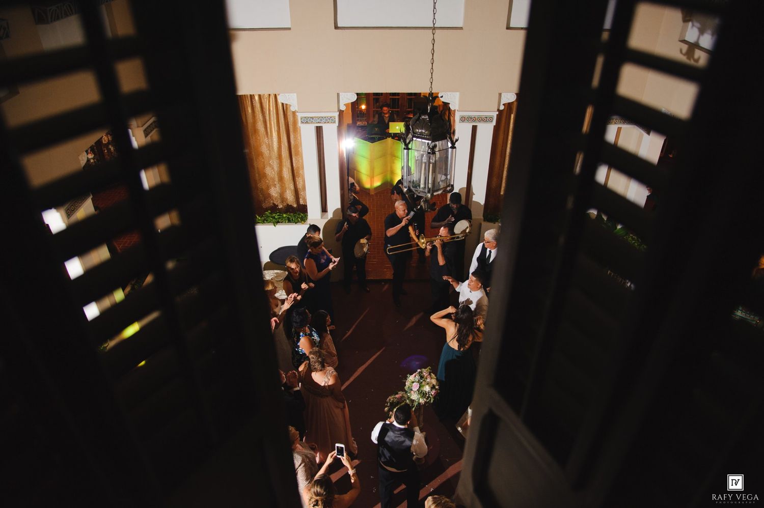 Aerial view of elegant indoor gathering with guests mingling in warmly lit venue, captured through decorative wooden shutters.