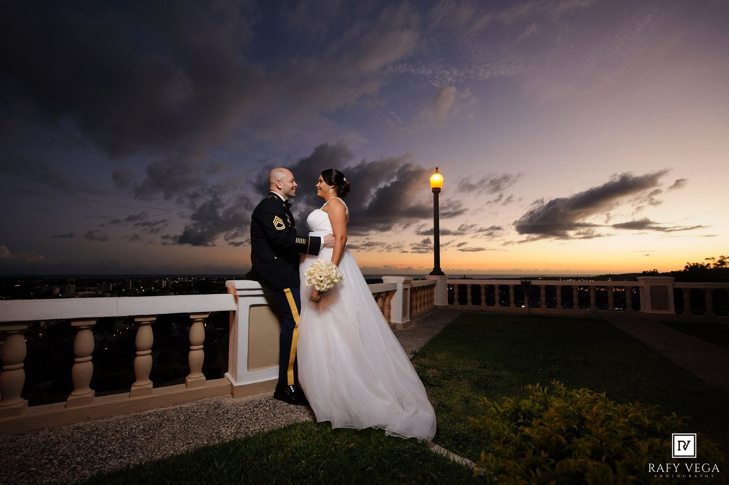 Military groom and bride pose romantically at sunset on a balcony, with a glowing streetlamp and dramatic sky backdrop.