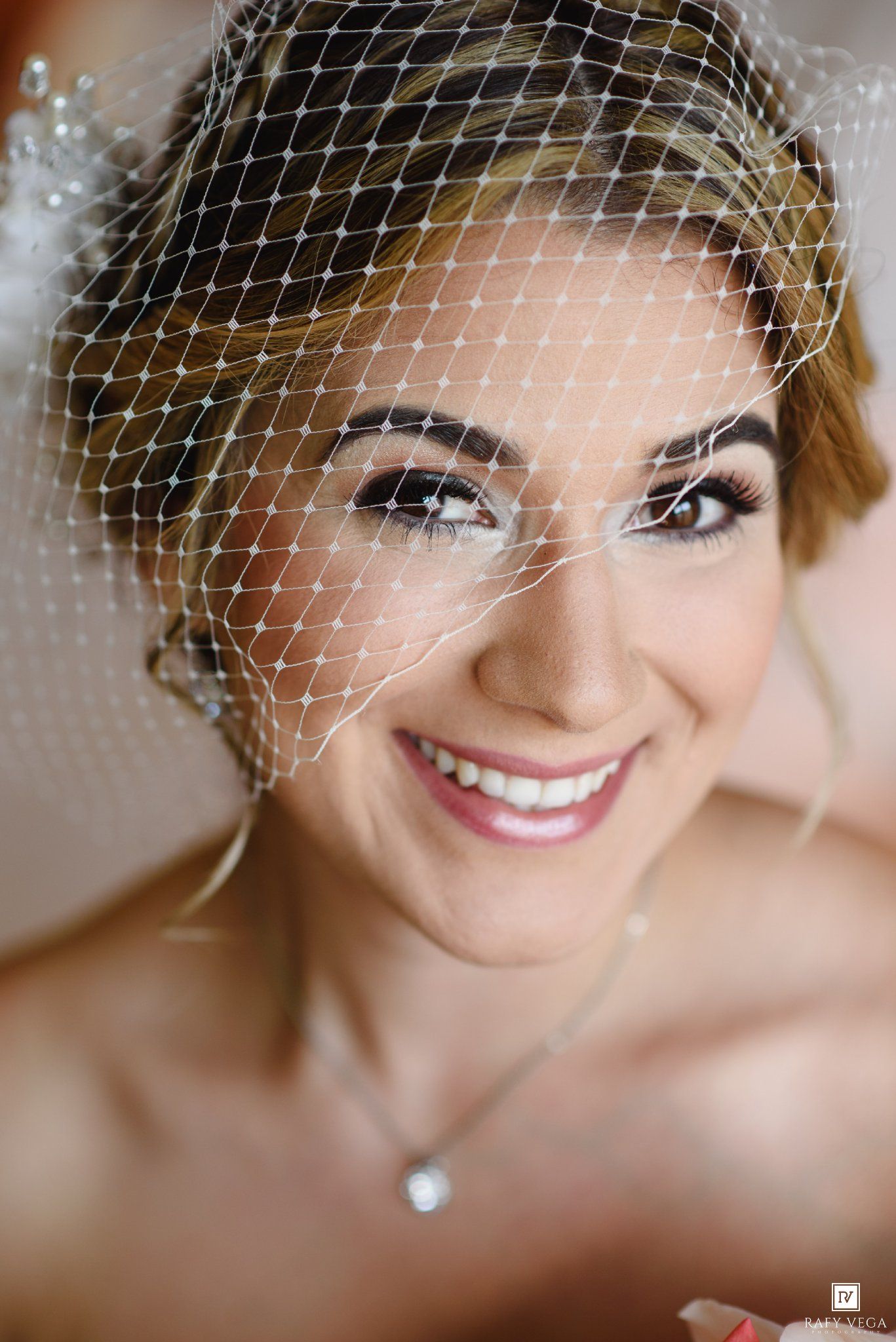 Smiling bride wearing a birdcage veil and delicate necklace with elegant makeup on her wedding day.